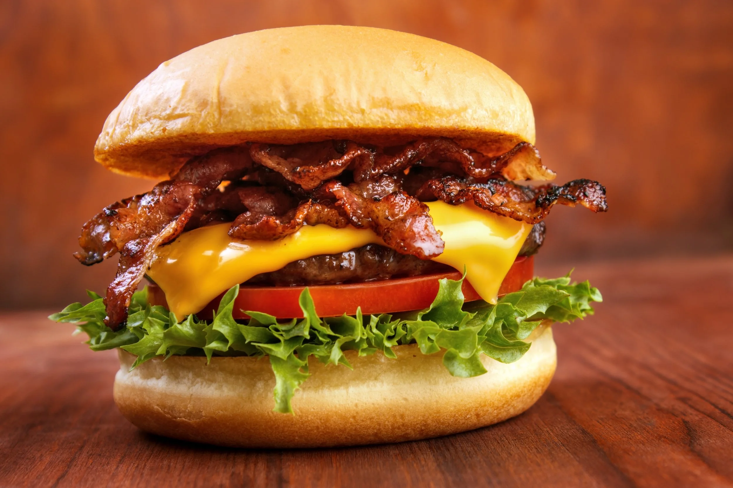 Close-up of a bacon cheeseburger with lettuce, tomato, cheese, beef patty, bacon, and a bun on a wooden surface.