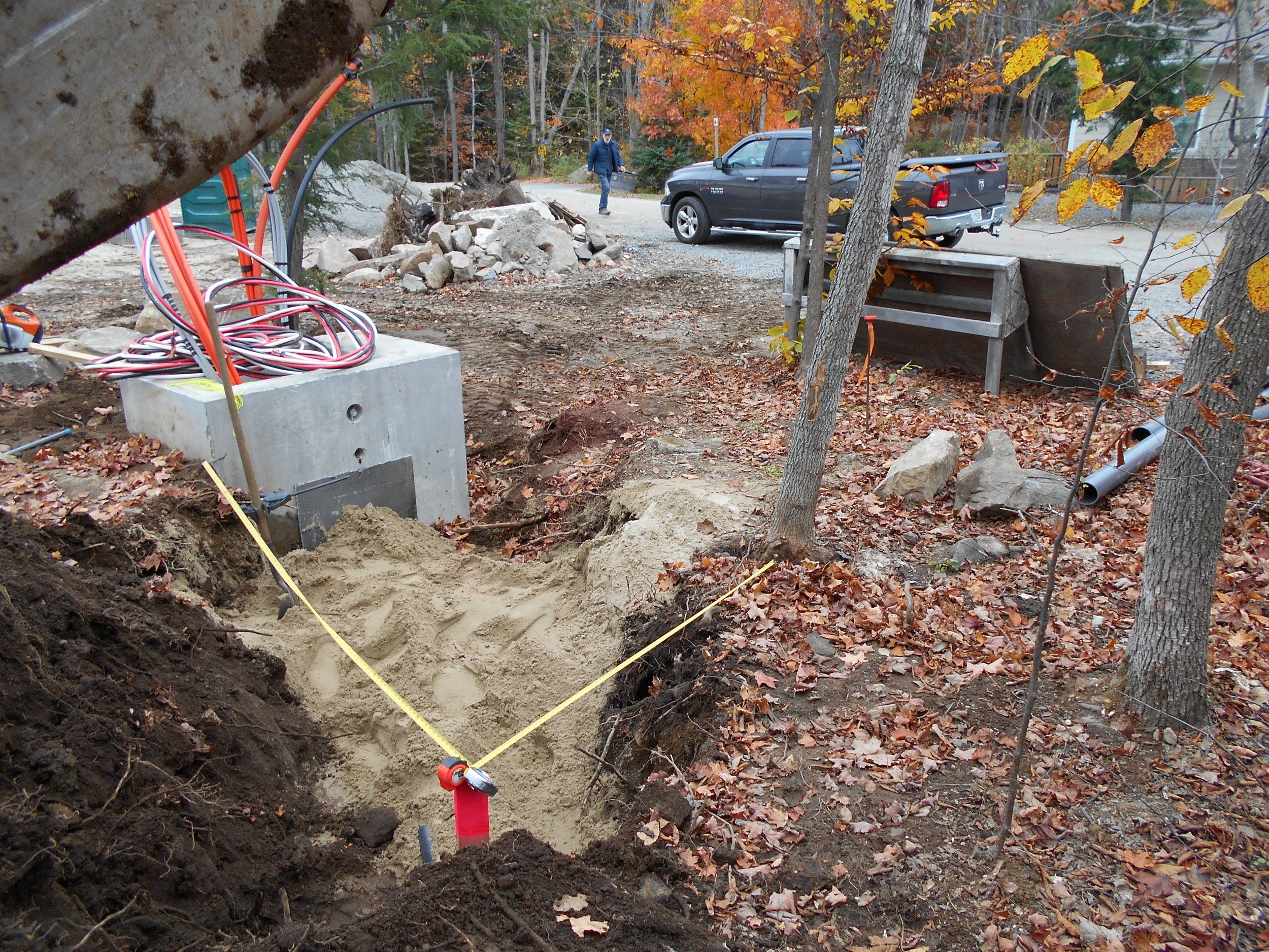 A construction or utility work site in a wooded area during fall, showing a large hole in the ground with exposed soil, utility pipes, and colorful autumn leaves on the ground. A person is seen walking in the background near a dark pickup truck parke