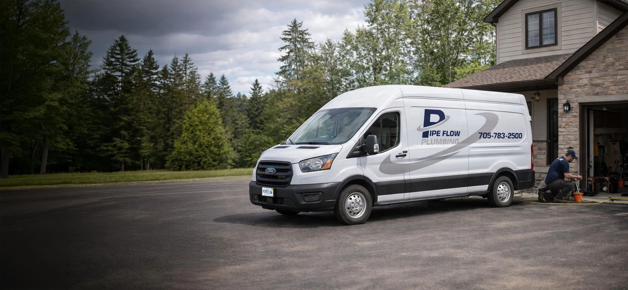 A plumber kneeling next to a white plumbing service van parked outside a house, connecting a hose to the house's outdoor faucet on a driveway surrounded by trees and shrubs.