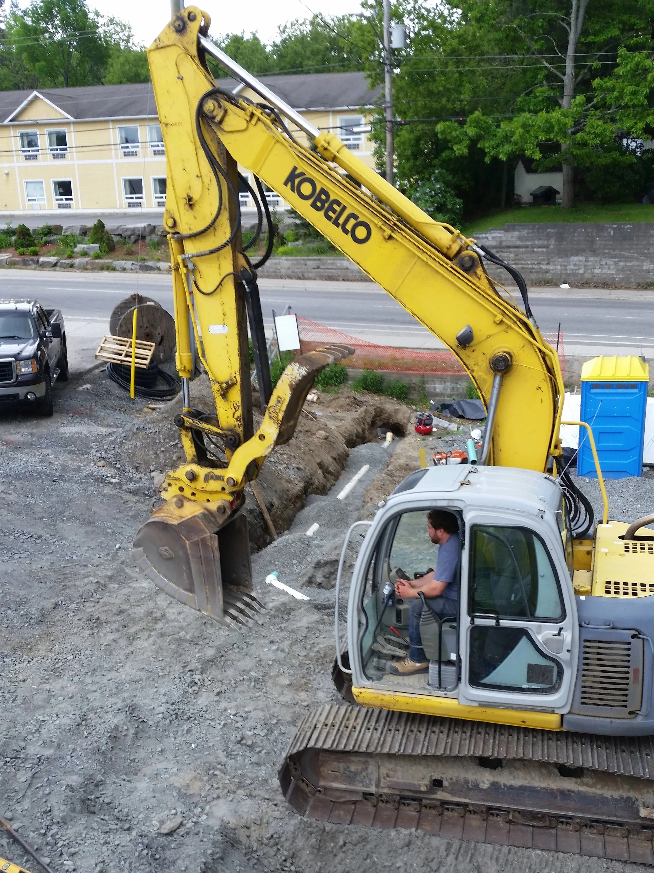 A construction site with a yellow Kobelco excavator digging a trench on a dirt surface. A man is seated in the excavator's cab, operating it. In the background, there are cars parked, a building with yellow siding, green trees, and power lines.