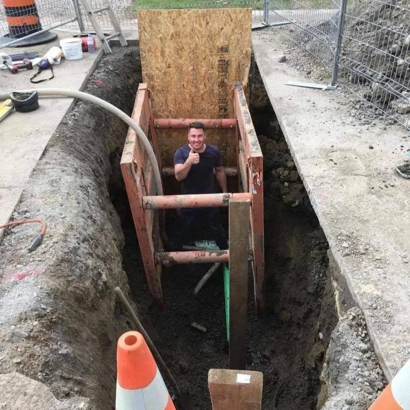 A man standing inside a construction excavation, giving a thumbs-up, surrounded by construction tools and materials.