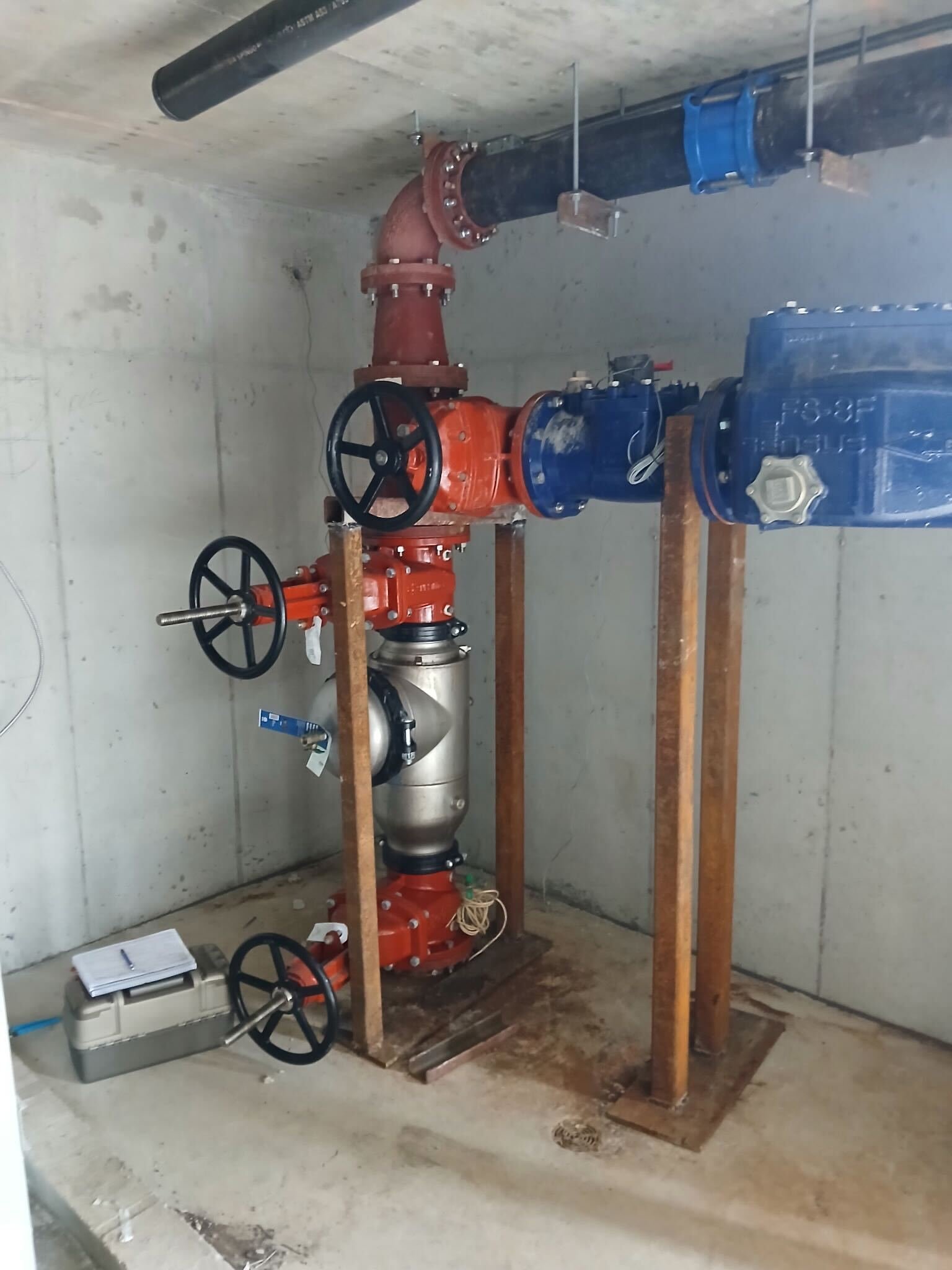An unfinished room with plumbing equipment, including large pipes, valves, a pump, and a filter, mounted on wooden supports. There's a notebook and some tools on the floor.