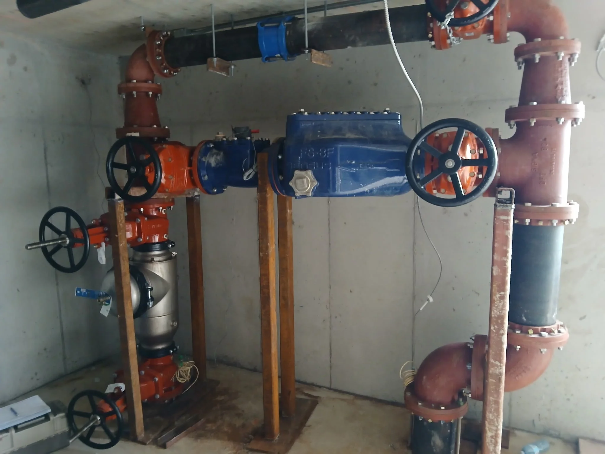 Underground plumbing system with large black pipes, orange valves, and blue components mounted on a wooden frame in a concrete room.