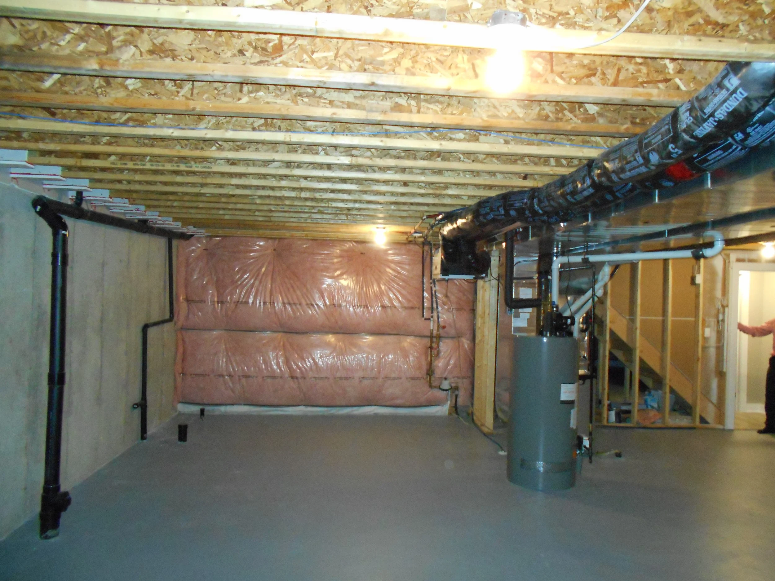 Indoor construction area with exposed wooden ceiling beams, black pipes, and a large pink insulation-covered wall in the background.