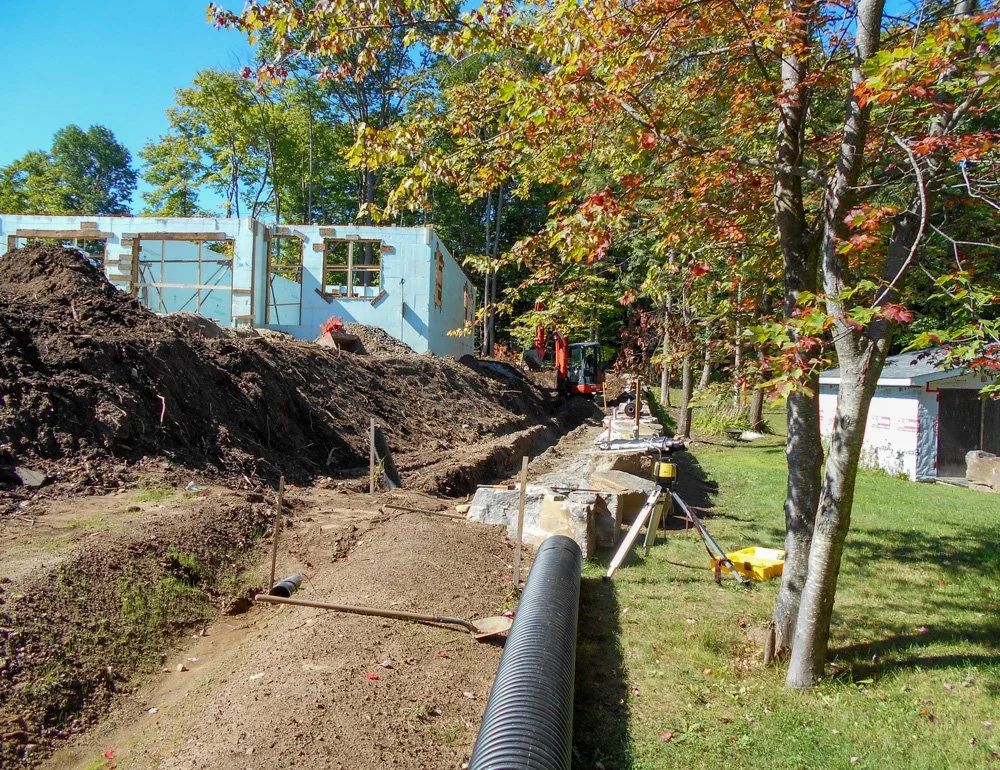 Construction site with excavated trench for plumbing, a partially built new home in the background, trees with fall foliage, and construction equipment.