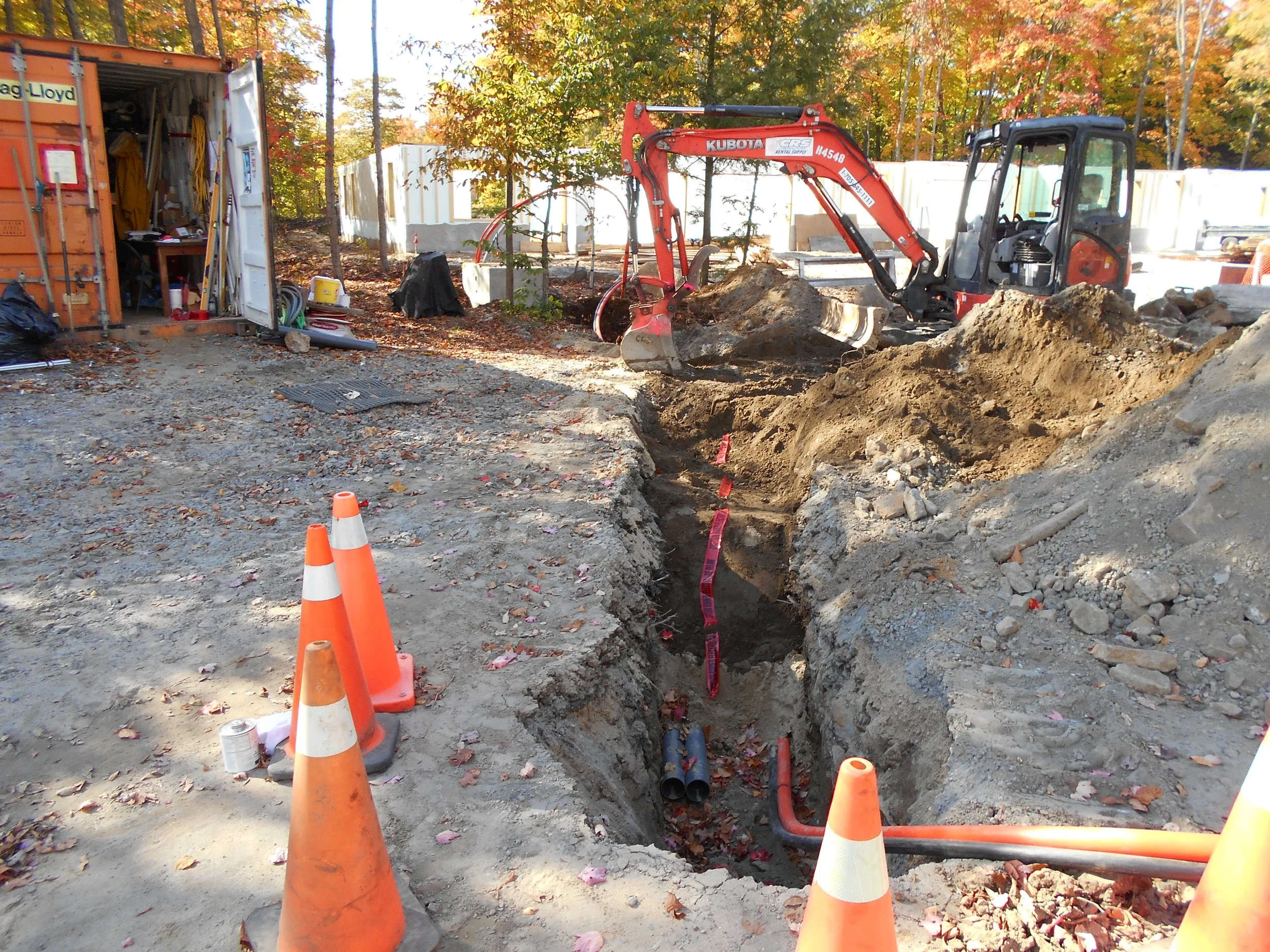 Construction site with an excavator digging a trench, orange and white traffic cones, a storage container with tools inside, and autumn trees in the background.
