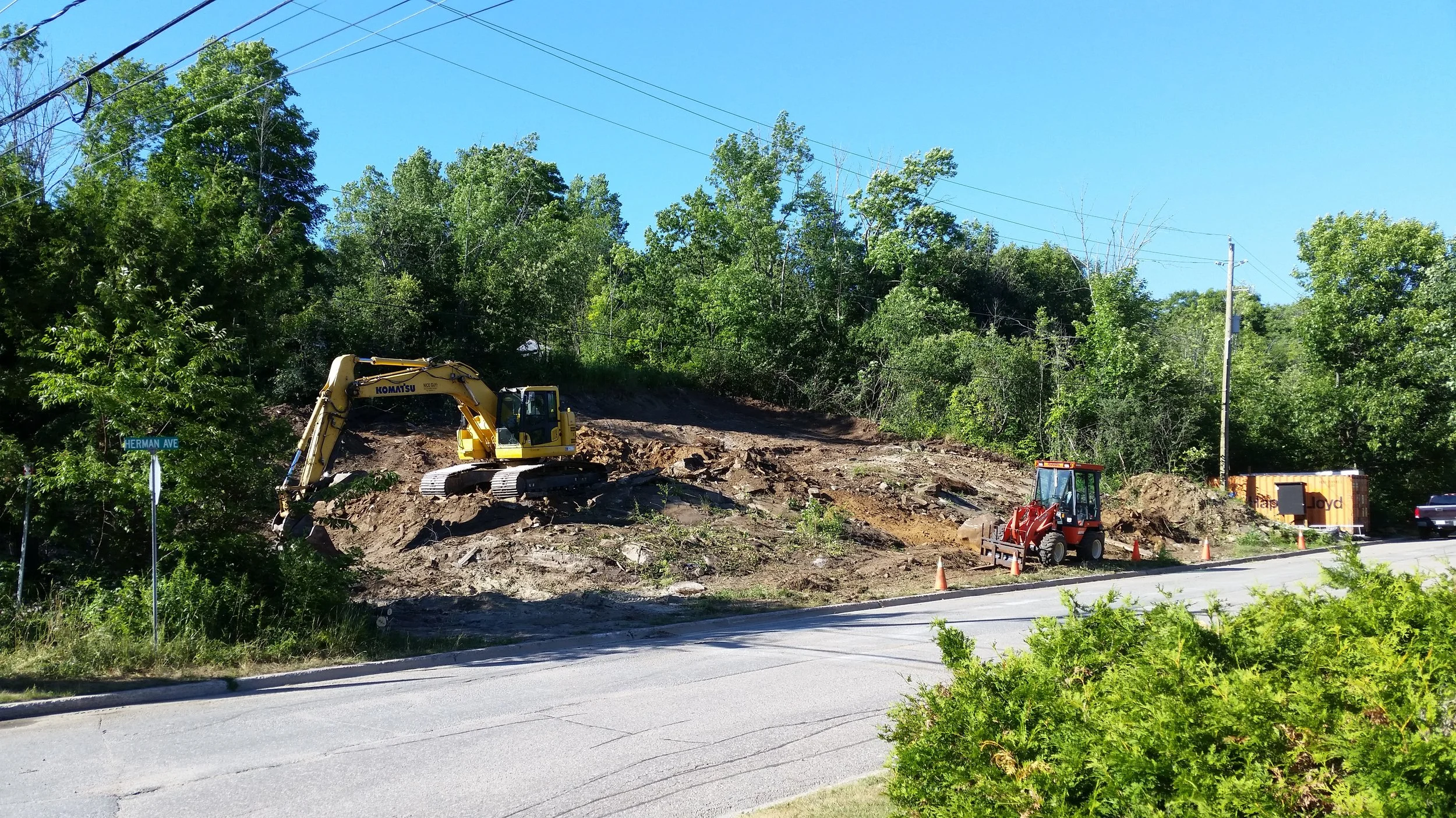 Construction site with a yellow excavator working on a hillside, a red tractor, and orange cones near Herman Avenue, surrounded by green trees and clear blue sky.