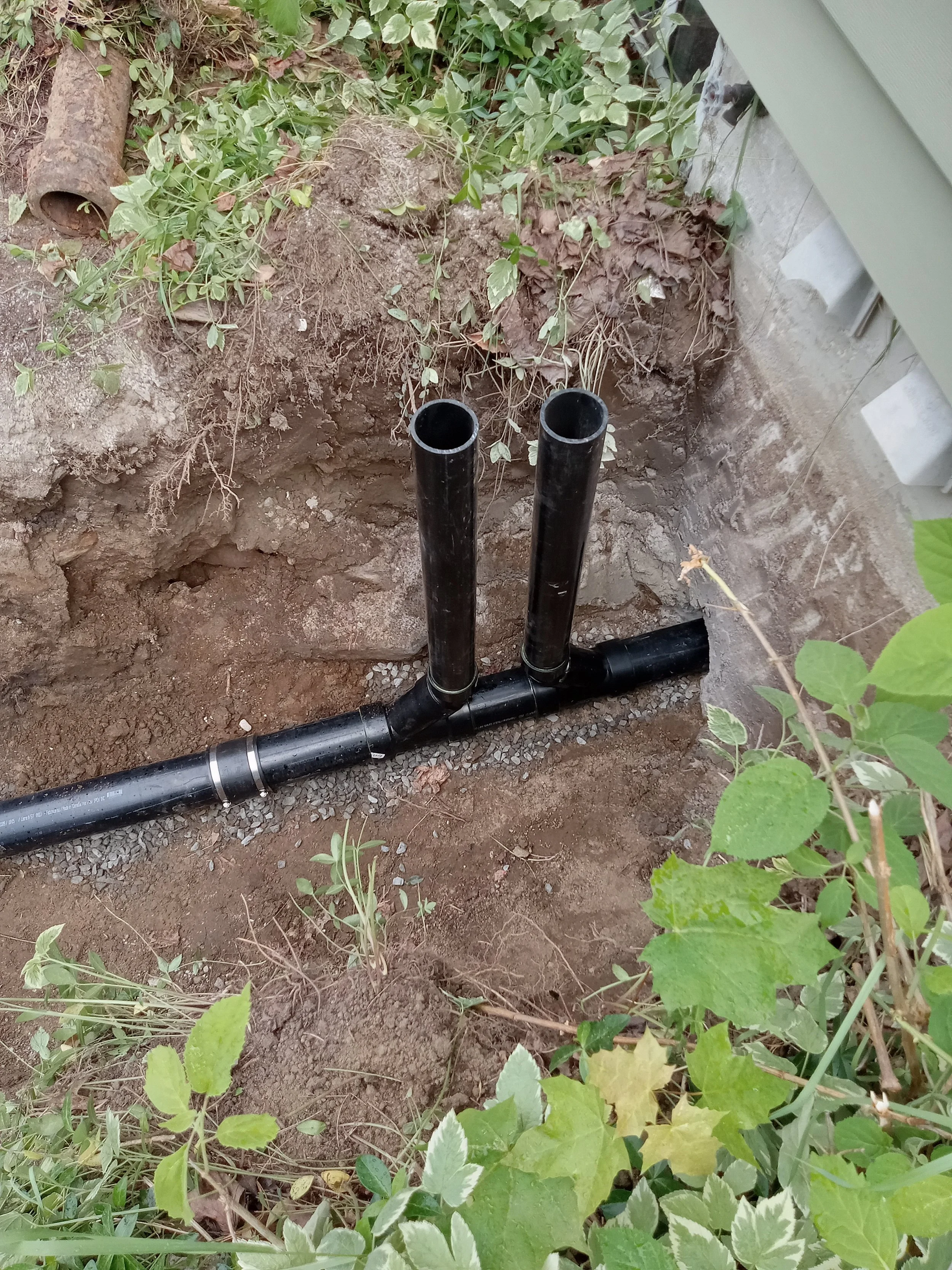 Black pipes are installed in the ground, emerging from the earth near a wall with some plants and weeds growing around.