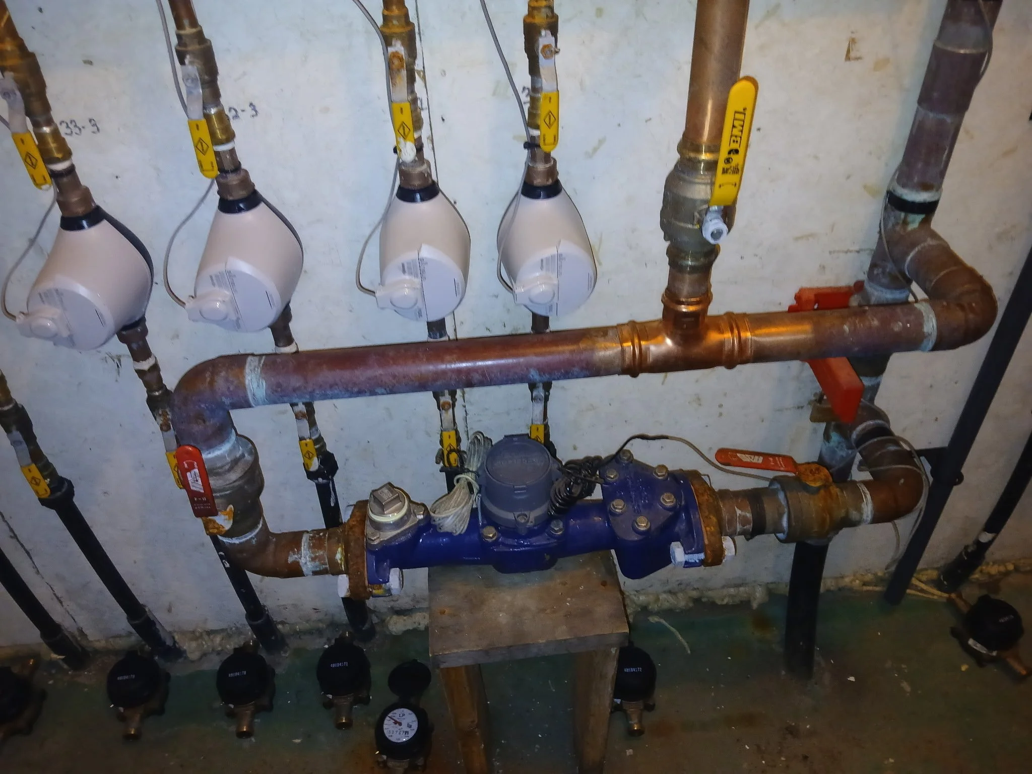 A plumbing setup with various pipes and valves, including a blue pump, electrical wiring, and white meters with black faces mounted on rusted pipes against a wall.