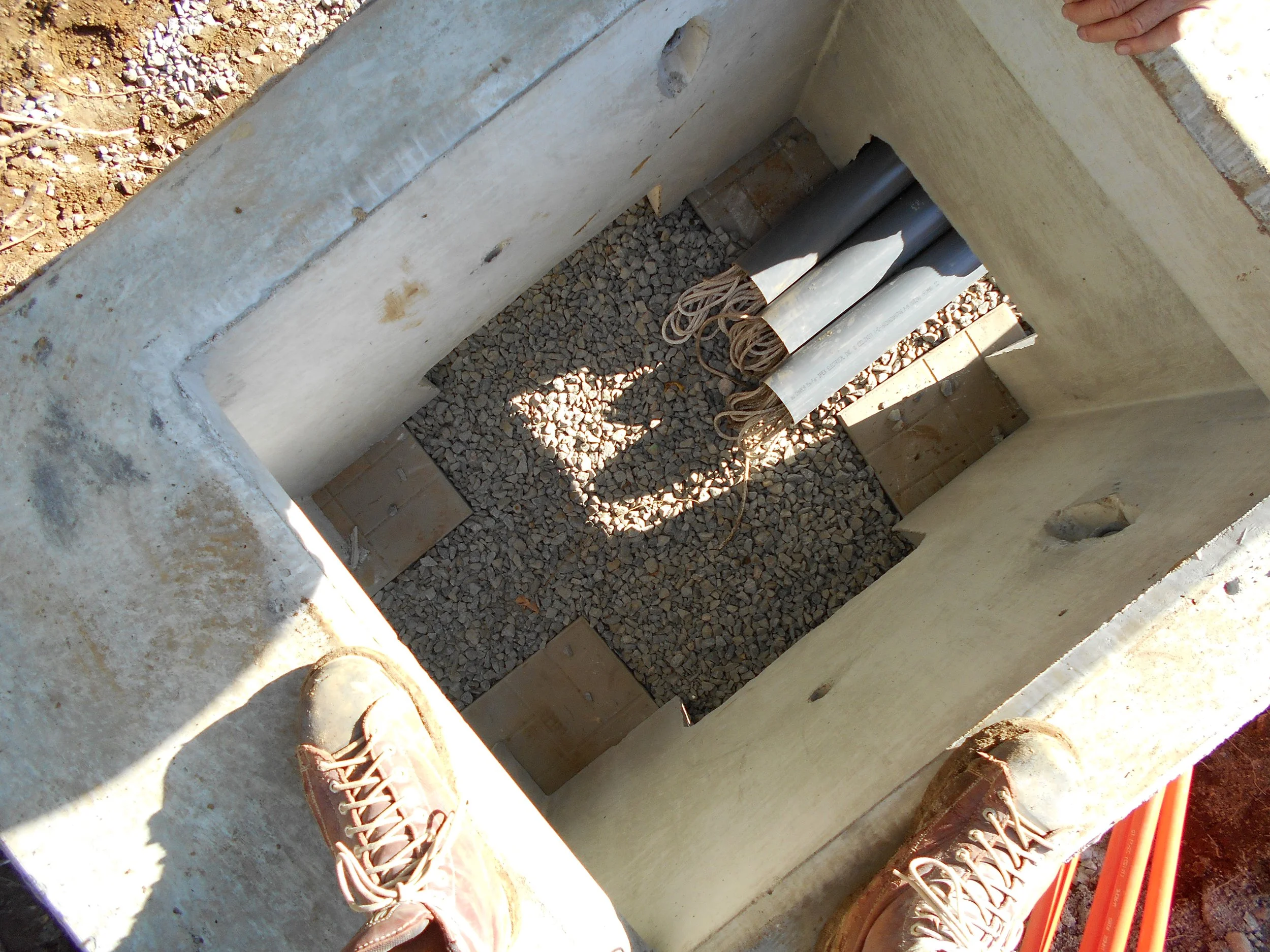 Construction worker's feet in brown work boots standing on a concrete foundation at a construction site, with electrical wires and pipes installed inside a small concrete structure.