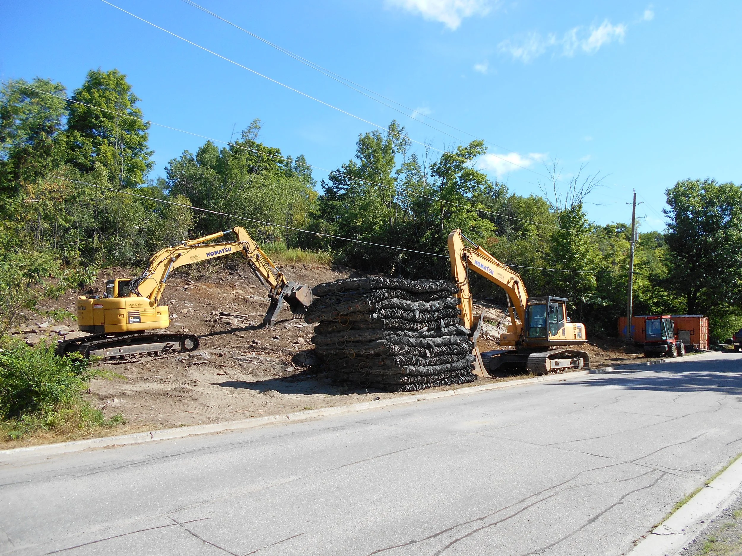 Construction site with two yellow excavators working on a hillside, with a large stack of black pipes or cables in the foreground, a row of trees in the background, and a blue sky with some clouds.