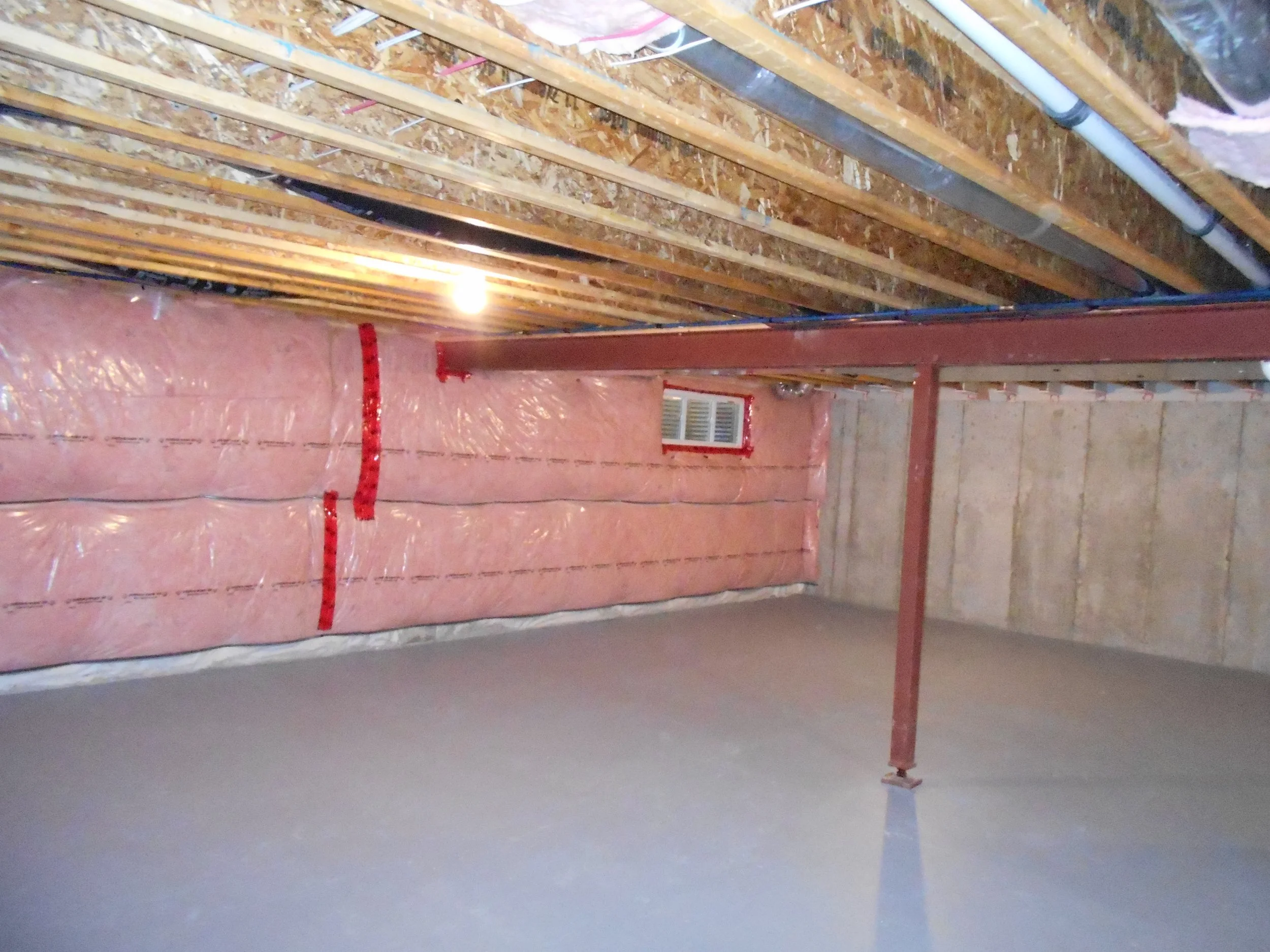 Interior view of a basement with exposed wooden ceiling beams, ductwork, electrical wiring, pink insulation, and a small vent window on the wall.