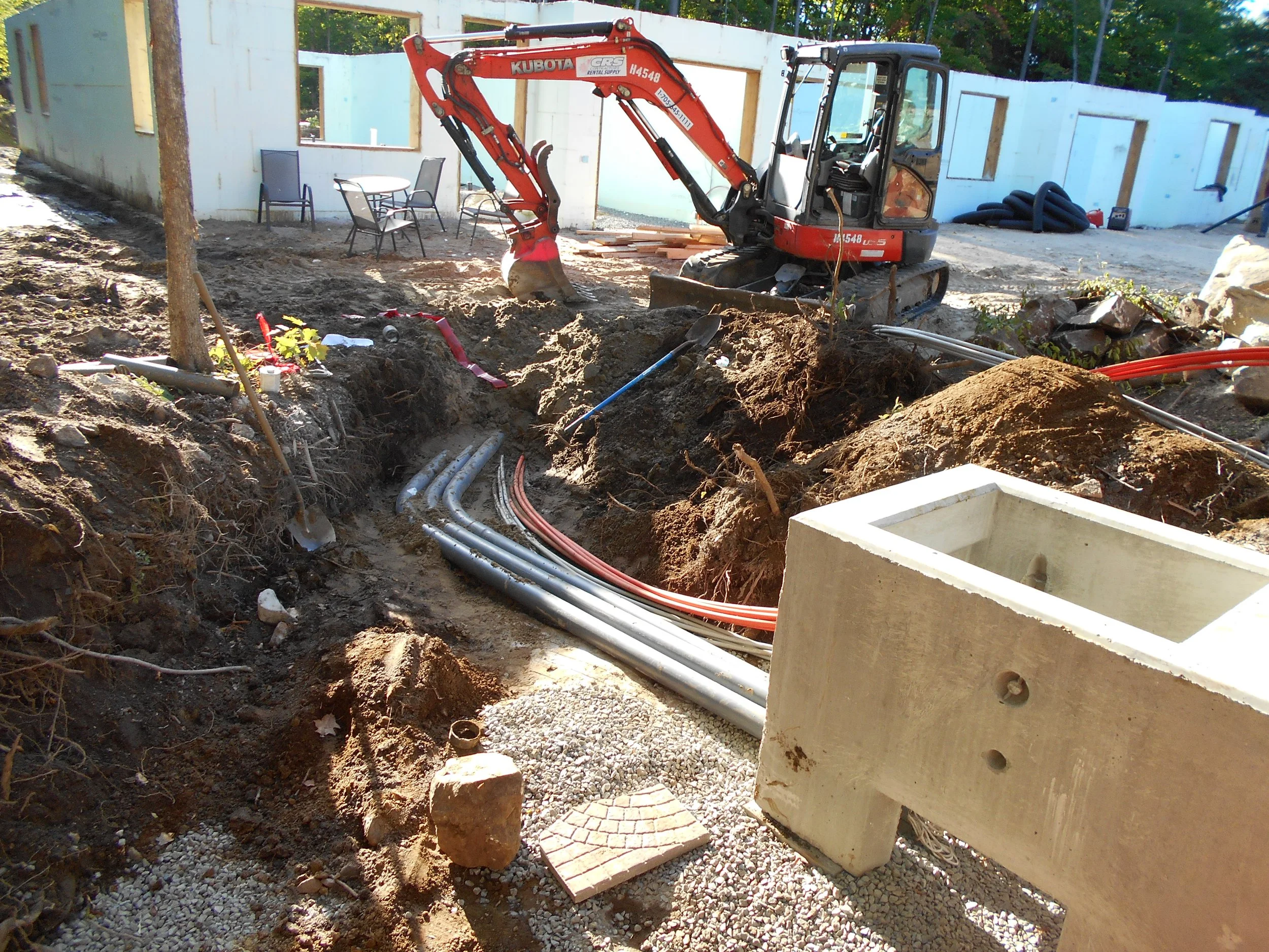 Construction site with a small excavator digging in the dirt, exposed plumbing pipes, a concrete structure, and a partially built white house in the background.