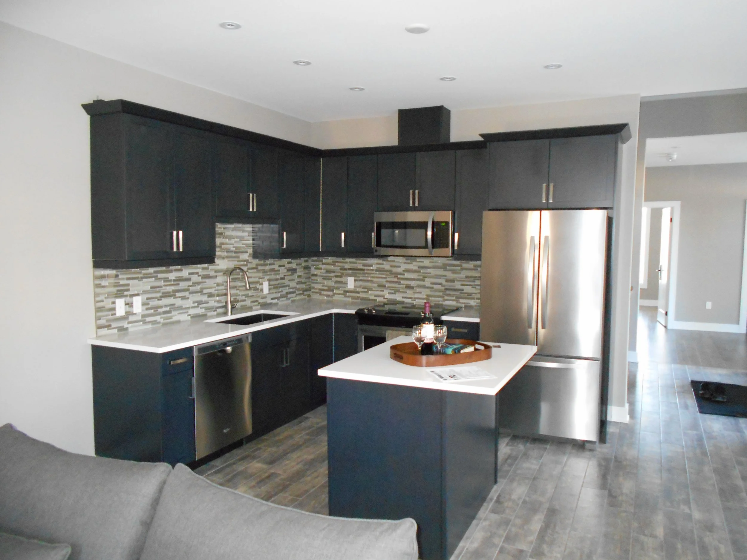 A modern kitchen with black cabinets, a gray tiled backsplash, and stainless steel appliances, including a refrigerator and microwave. There is a kitchen island with a white countertop, a tray with some glasses, and a sink with a high arch faucet. Th