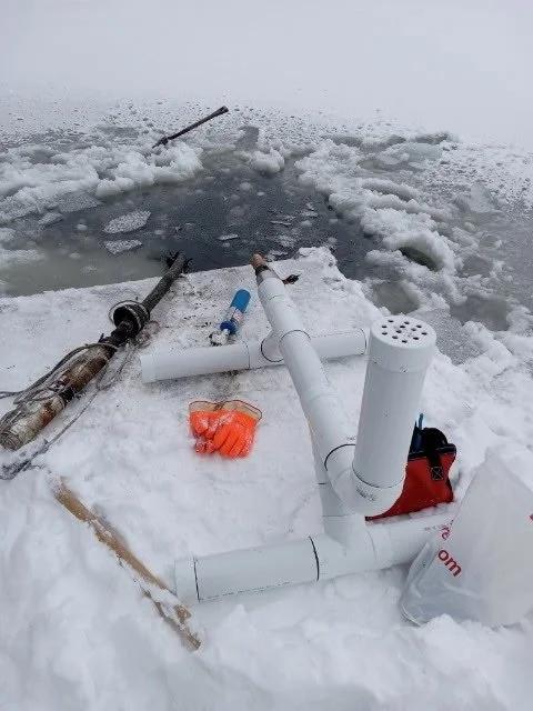 Ice fishing setup with a hole drilled in the ice, a fishing pole, and equipment on snow in a frozen lake.