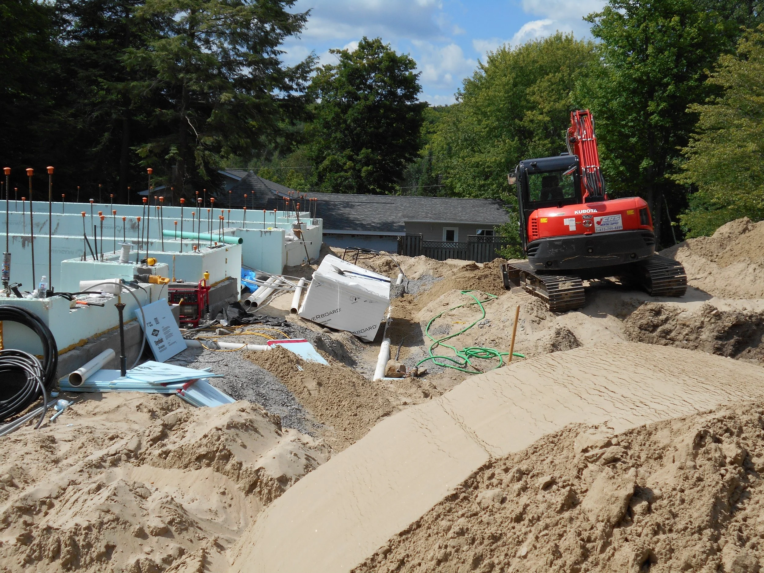 Construction site with a small red excavator working on the dirt. There are pipes, tools, and construction materials scattered around. Trees and a house are visible in the background.