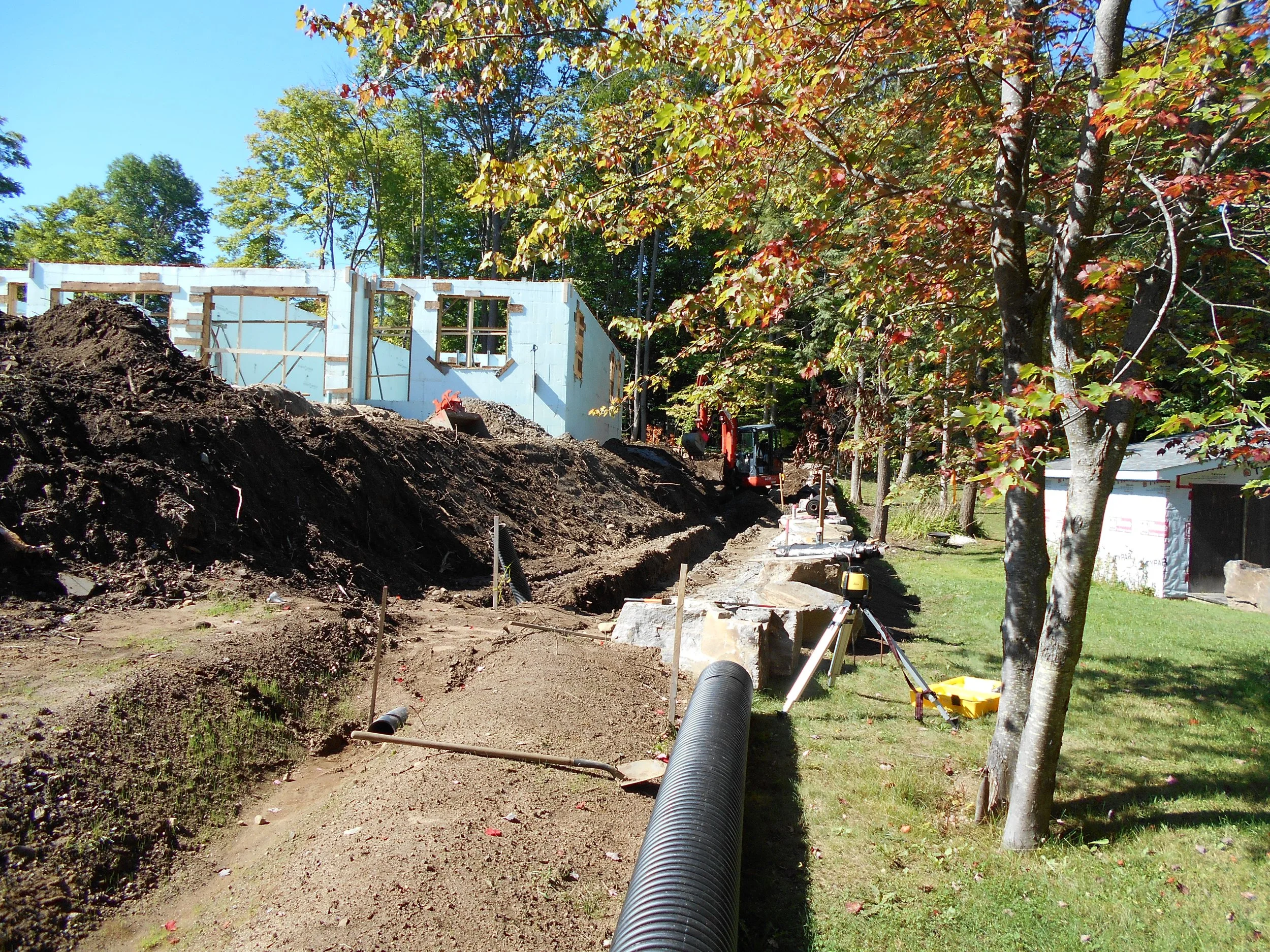 Construction site with partially built wall, dirt, and construction equipment surrounded by trees with autumn leaves.