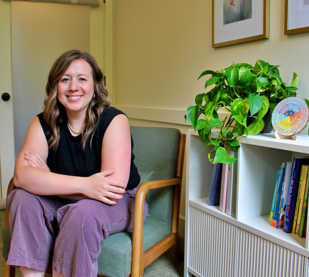 A woman with shoulder-length wavy hair, wearing a black sleeveless top and striped pants, sitting on a green chair with her arms crossed, smiling. She is in a room with framed artwork on the wall, next to a white shelf with books and a green leafy plant.