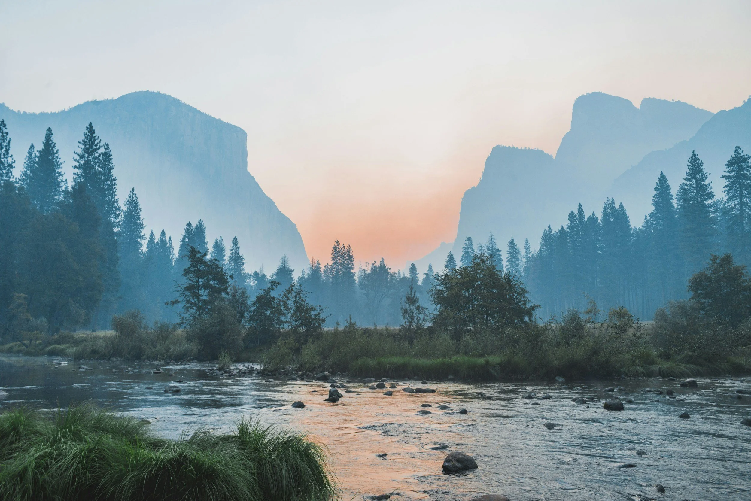 A serene landscape featuring a river flowing through a forest with tall trees and distant mountains at sunset or sunrise.