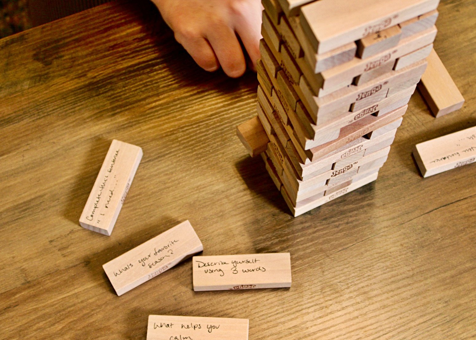 A Jenga tower on a wooden table with questions written on the wooden pieces. One wooden piece says, 'Describe yourself using 3 words,' another says, 'What's your favorite season?,' and others have questions like 'What helps you calm?'