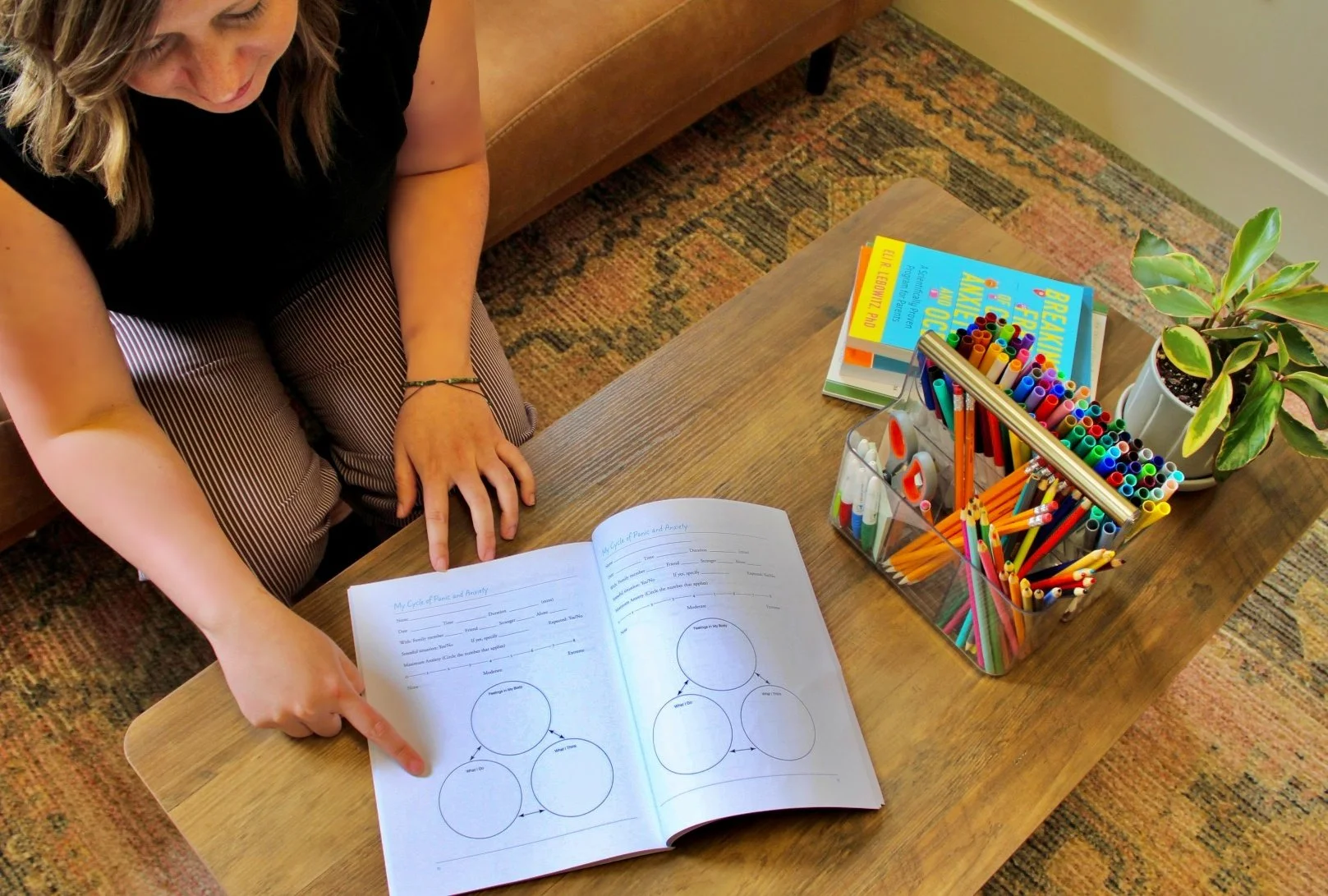 A woman sitting at a wooden table with a book open, pointing to a diagram. The table has a box of colorful markers, highlighters, and pens, a stack of books, and a potted plant. The area has a colorful carpet and a brown sofa in the background.
