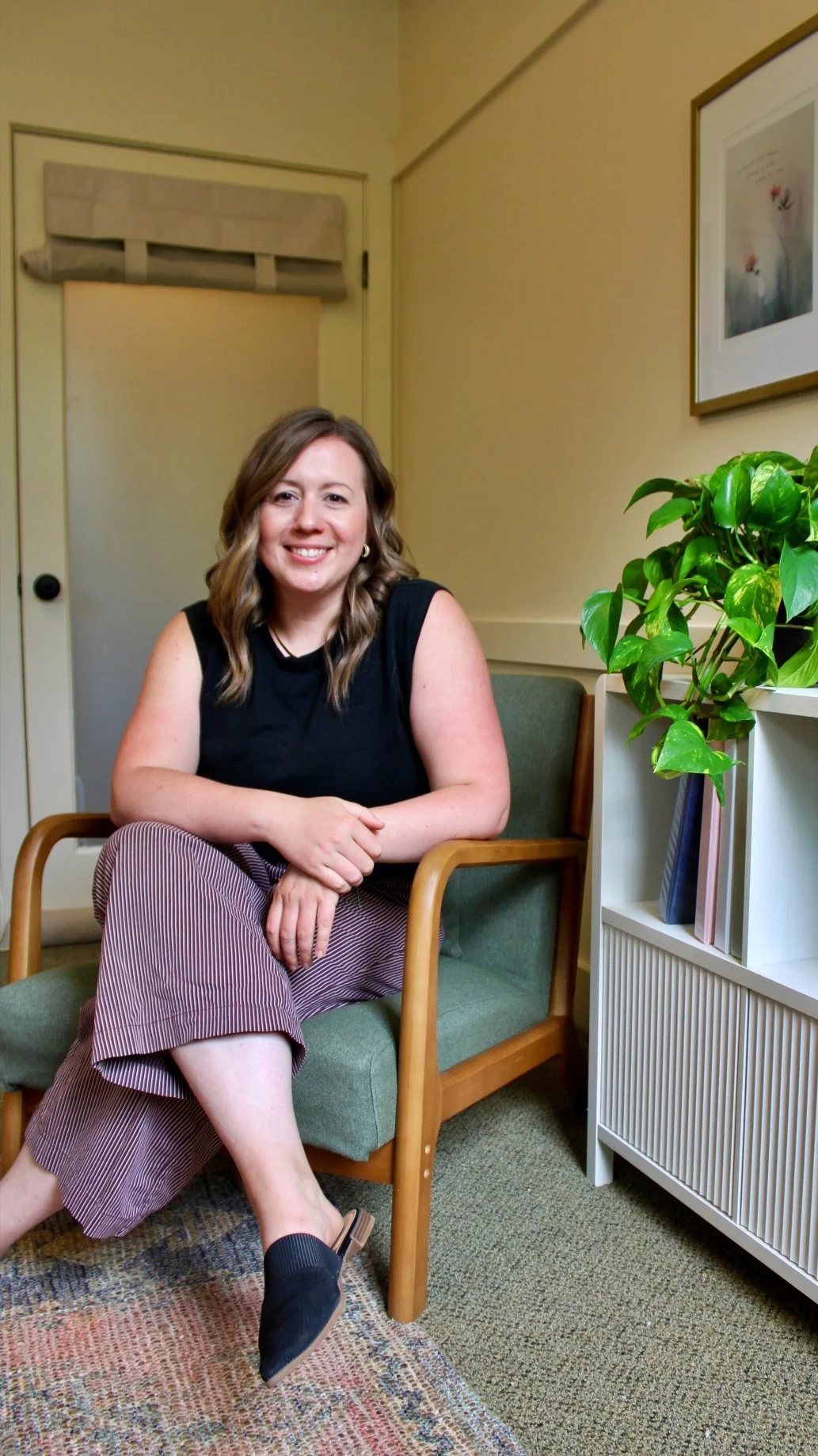 A smiling woman with shoulder-length brown hair sitting on a green upholstered chair with wooden arms in a cozy room. She is wearing a black sleeveless top, brown striped pants, and black slip-on shoes, with her hands resting on her lap. There is a white bookshelf with books and a plant on her right, and a framed picture hanging on the beige wall behind her. A potted green leafy plant is on top of the bookshelf, and a beige door with a rounded top is visible in the background.