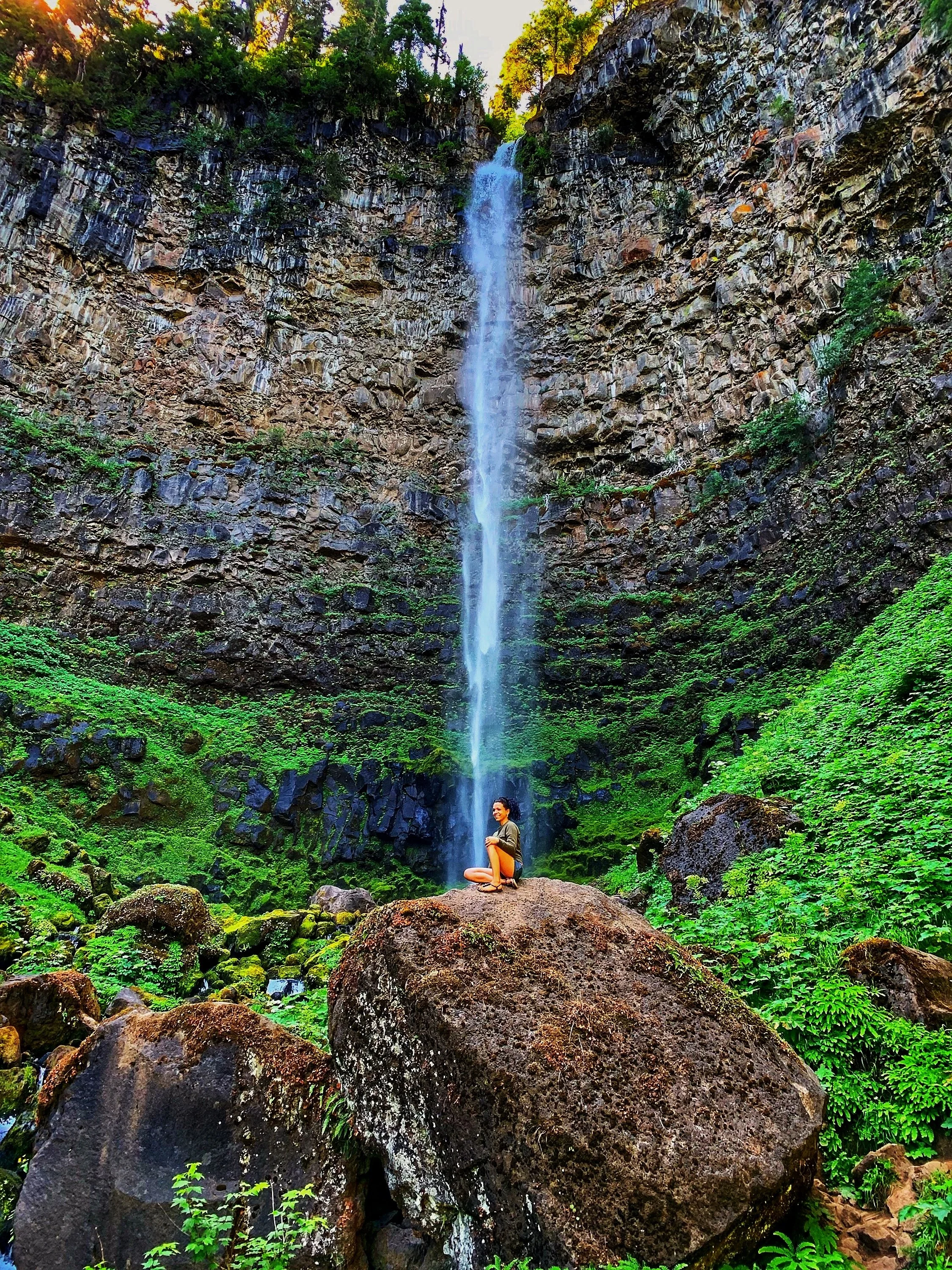 A woman sitting on a large rock in front of a waterfall surrounded by green foliage and moss-covered rocks.