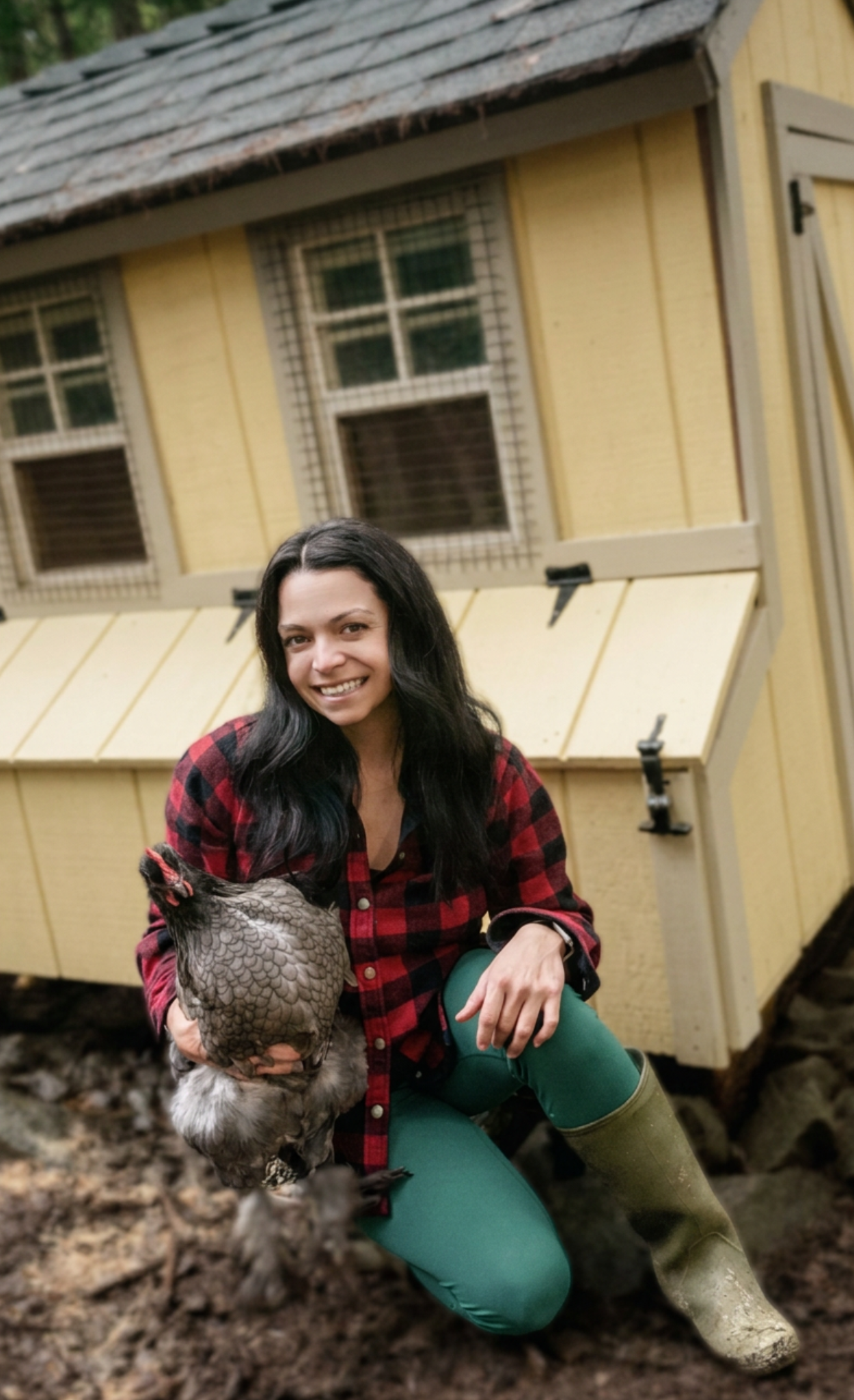 Woman kneeling on the ground outdoors, holding a chicken, in front of a yellow shed with barred windows, dressed in a red plaid shirt, green pants, and rubber boots.