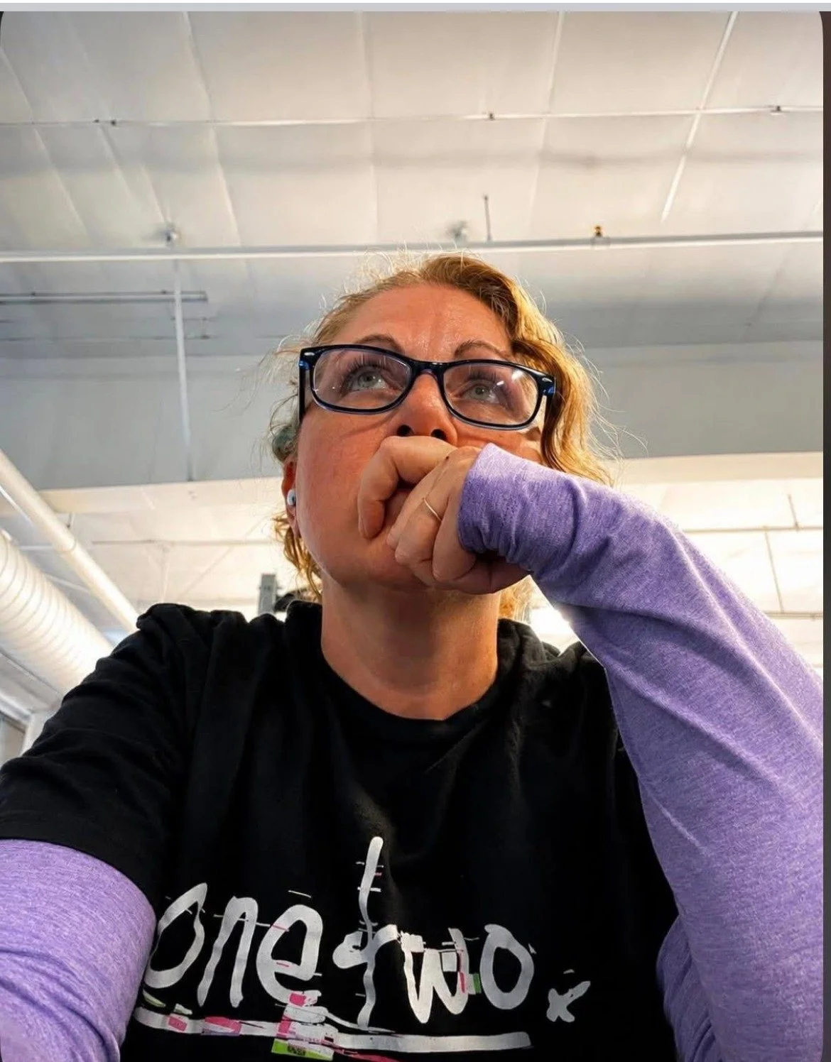 A woman with curly blonde hair and glasses sitting indoors, resting her chin on her hand, wearing a black t-shirt and purple long-sleeve shirt, looking thoughtfully upward.