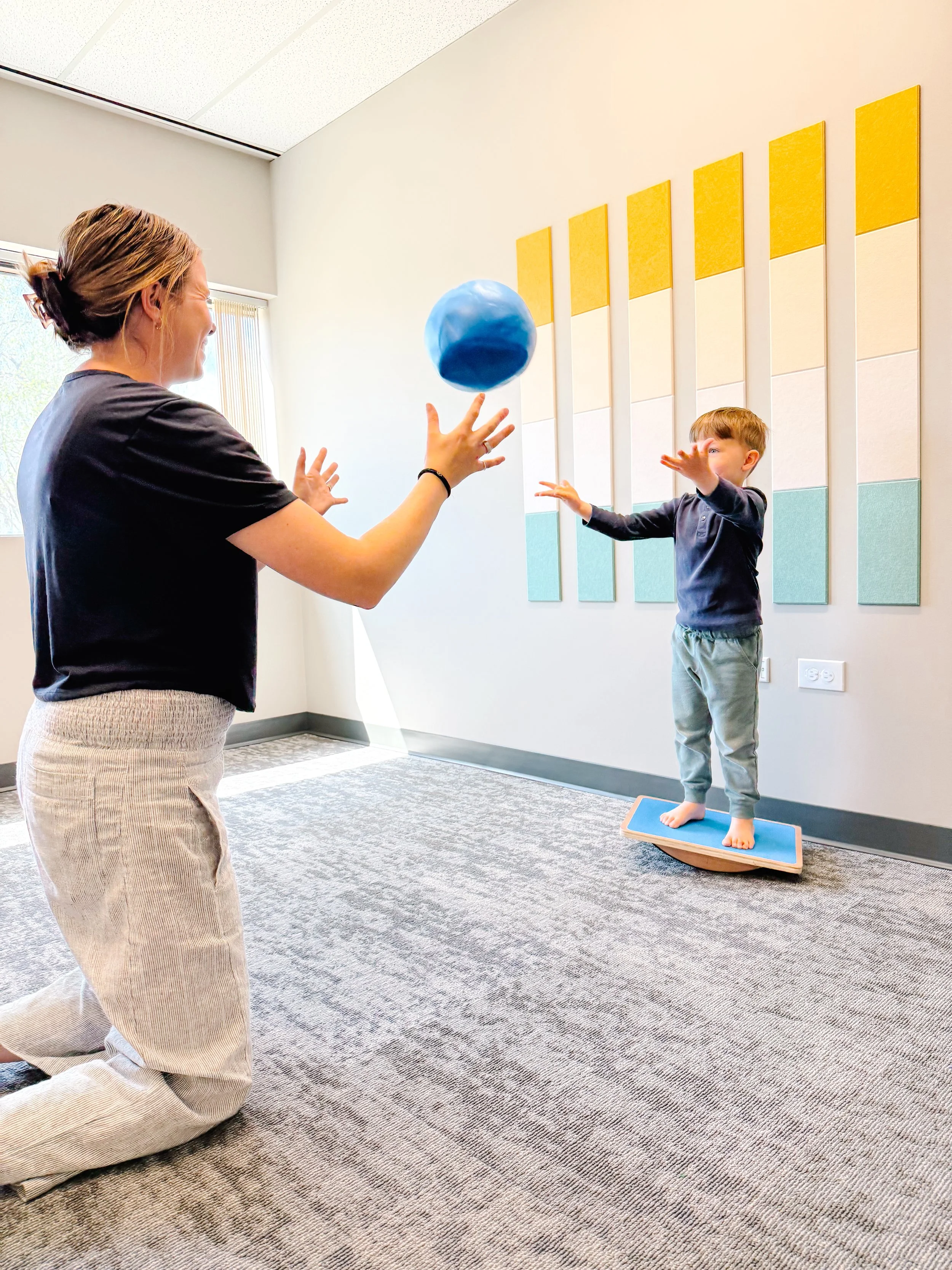 therapist kneeling and child standing on balance beam throwing ball