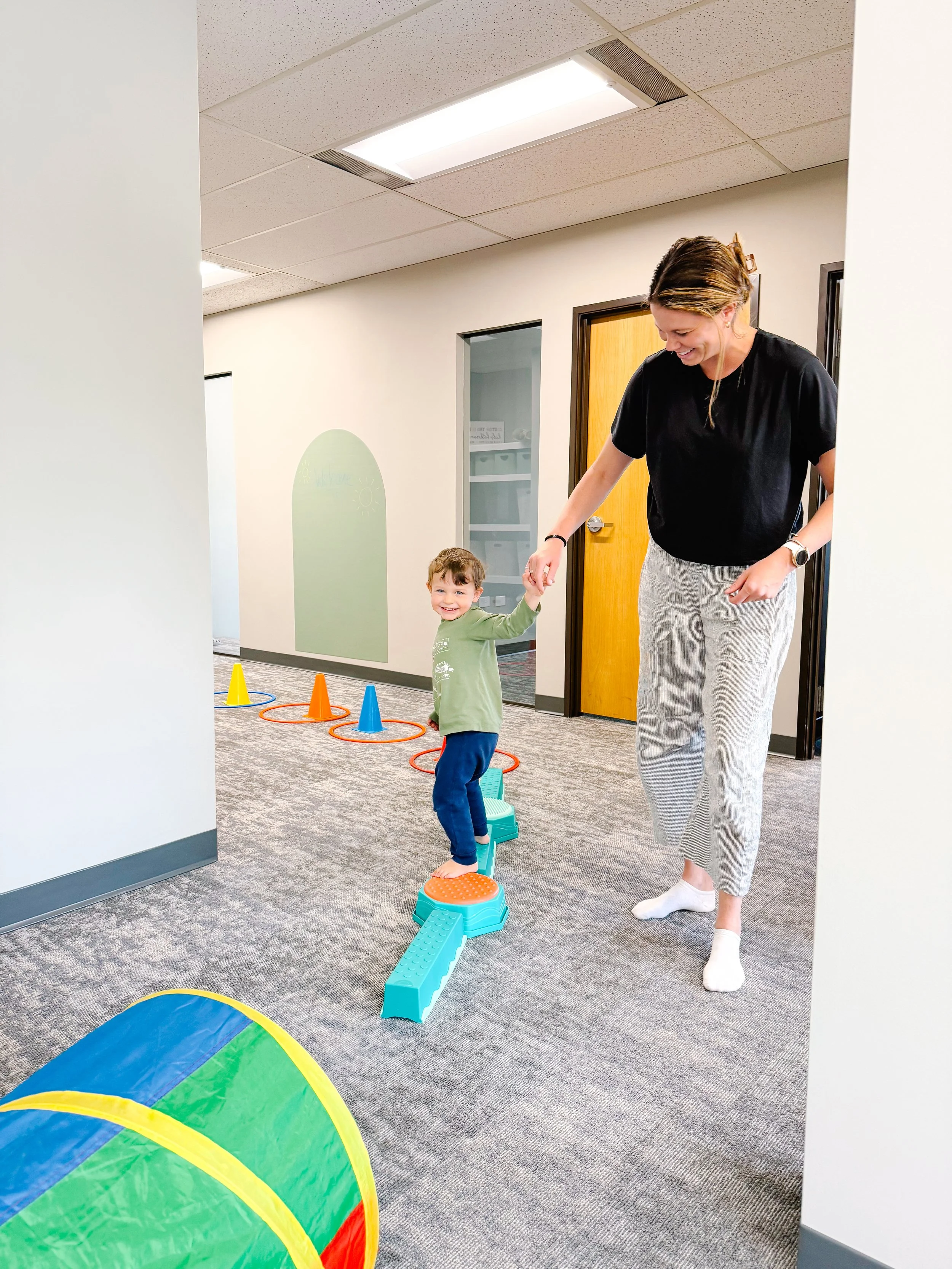 child doing obstacle course on balance beam holding therapist hand, both smiling