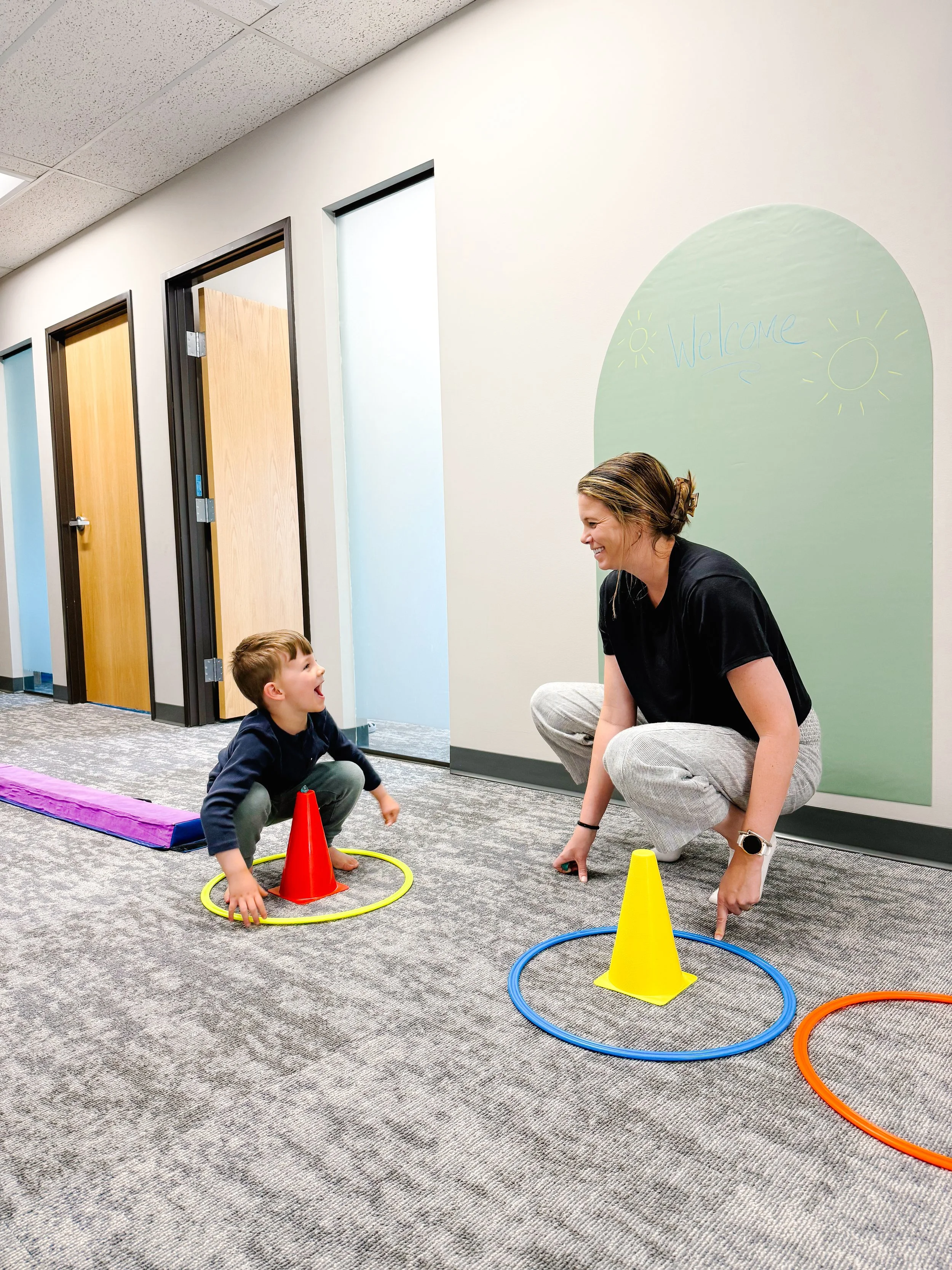 female therapist in a squat postion smiling looking a young boy in same position while doing an obstacle course