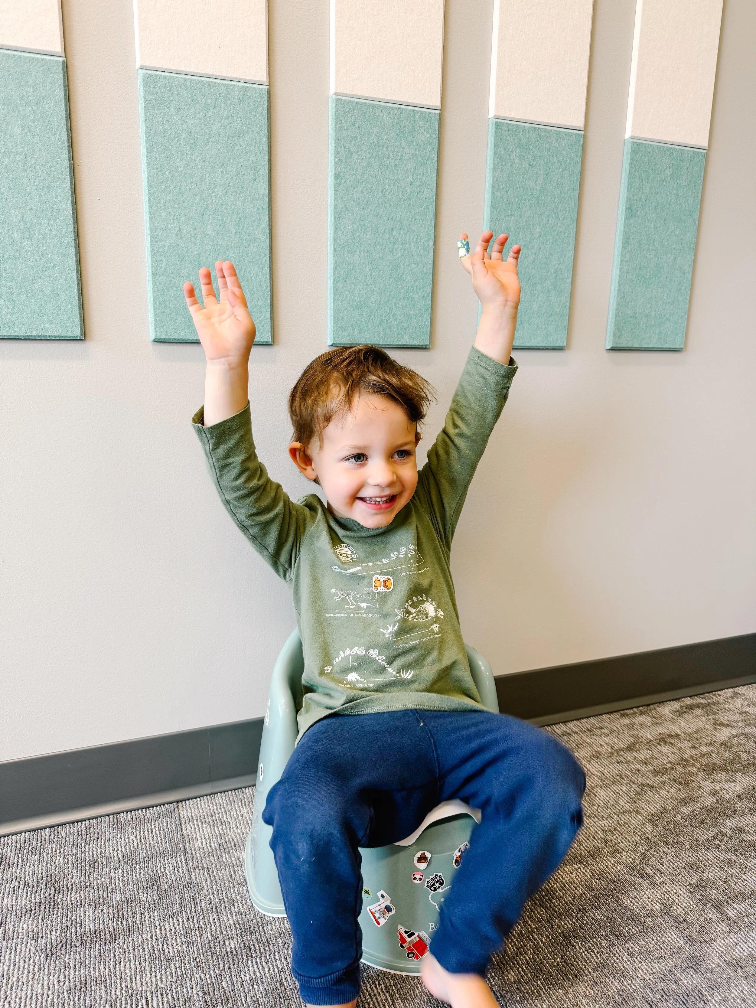 young boy sitting on small floor potty with hands in air happy