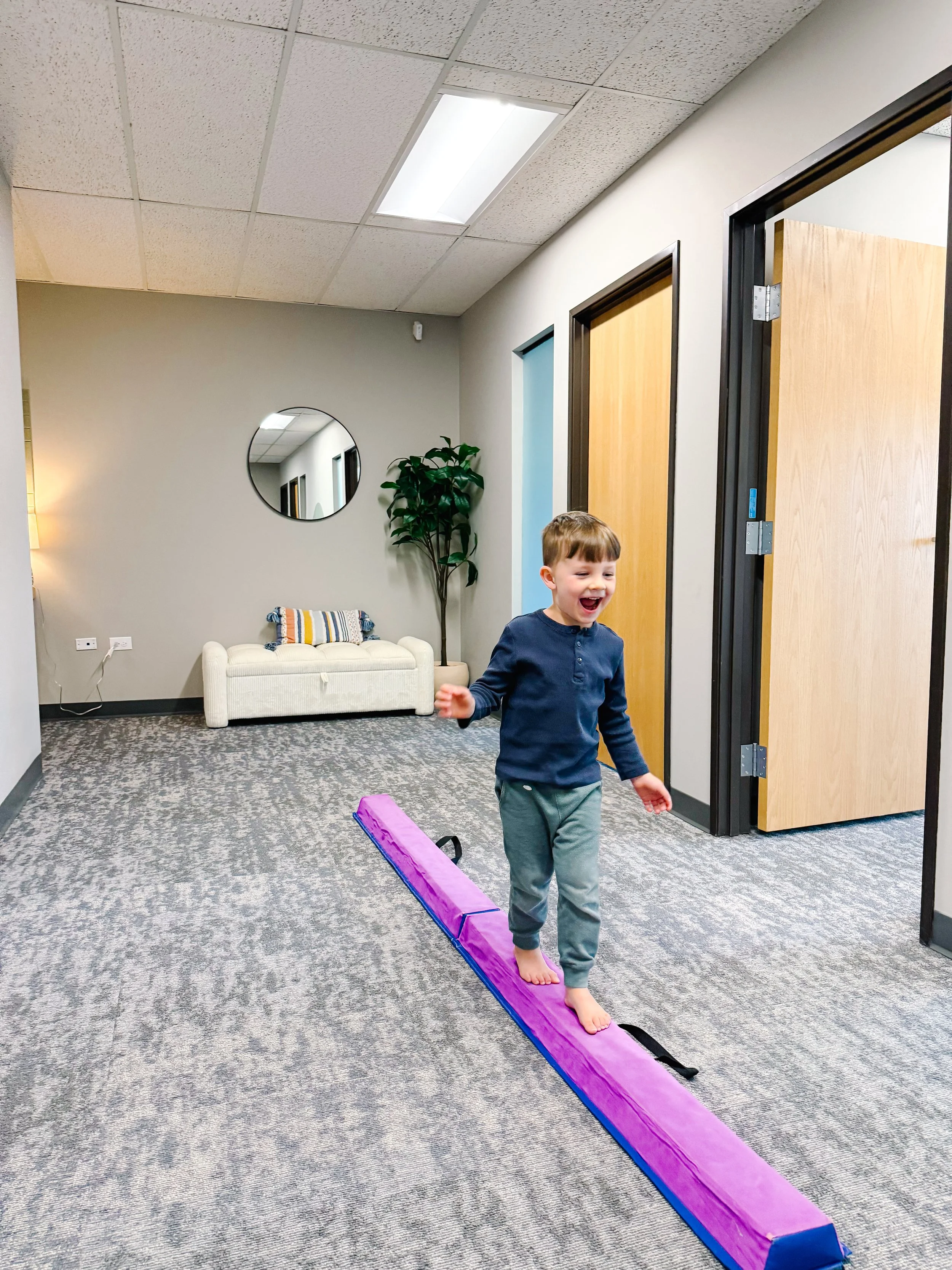 boy smiling, walking across balance beam in therapy clinic