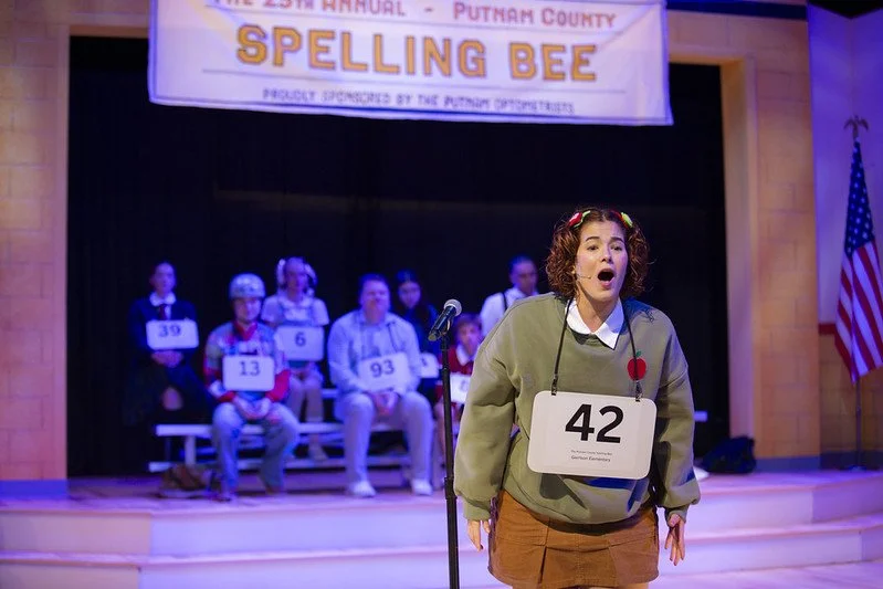 A girl with curly hair standing on stage at a spelling bee, wearing a number 42 on her shirt. In the background, several people with large numbers sit on stage, and a banner overhead reads 'The 25th Annual - Putnam County Spelling Bee'. An American f