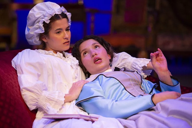 Two women dressed in historical or theatrical costumes sitting close together on a red sofa, with one woman resting her head on the other's chest. The scene appears to be from a play or historical reenactment.