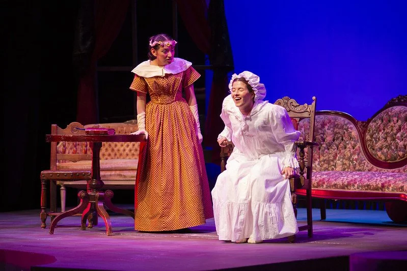 Two women in vintage costumes performing on stage, one standing and the other sitting, with period furniture and a blue backdrop.