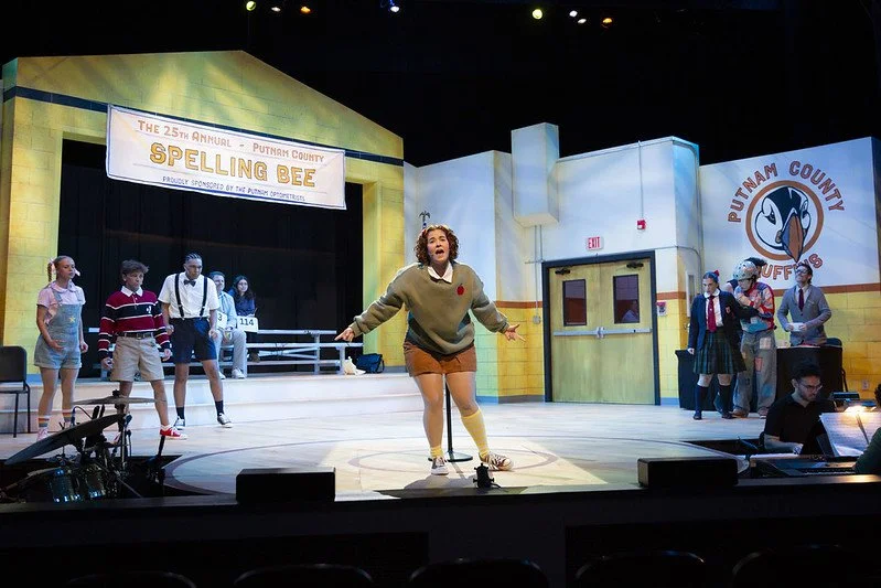 The stage set for a theatrical performance of a school play called 'Spelling Bee,' featuring performers in school costumes. A girl is at the center with others standing on either side, with a focused audience watching.