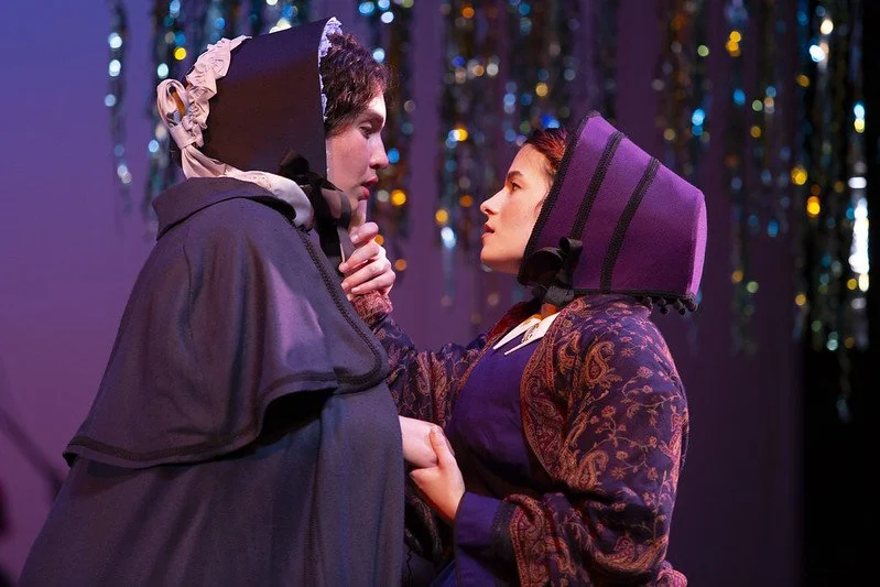 Two women in vintage costumes standing close, looking into each other's eyes in front of a colorful, reflective tinsel curtain stage background.