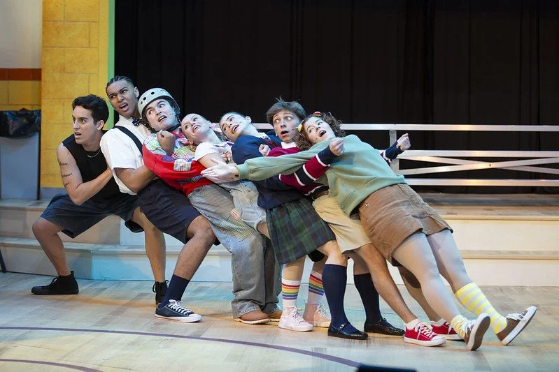 Group of children and teenagers on stage in a theatrical performance, leaning back in a line with exaggerated facial expressions and colorful costumes.