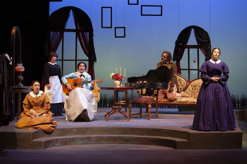 Stage scene with five women, some sitting and some standing, involved in a theatrical performance. One woman is playing guitar, another is seated beside her, and the others are standing, dressed in period costumes. The background features large arche