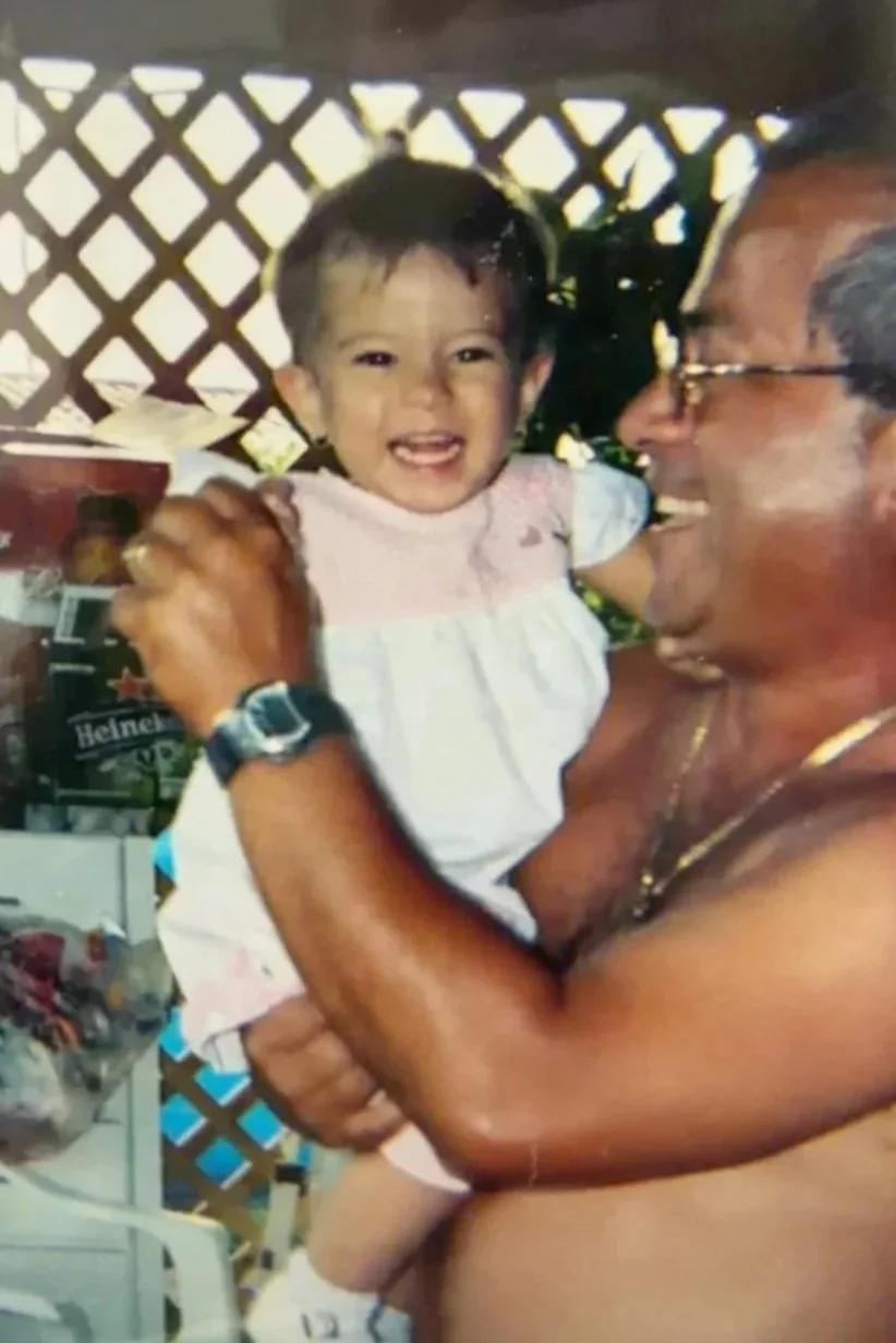 A man with glasses holding a young smiling girl with short dark hair, wearing a light pink and white dress, in an outdoor area with lattice fencing and various items in the background.