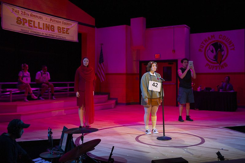 A young girl with a contest number 42 stands at a microphone on stage during a spelling bee in Putnam County. Two girls are sitting on the riser behind her, and a performer in a mask and red dress stands on the side. A man is taking a photo, and a wo