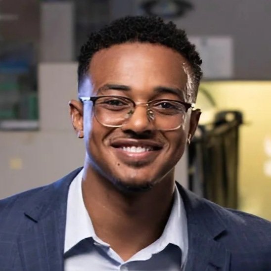 A young man with short curly hair, glasses, and a beard, smiling in a professional setting, wearing a suit and white shirt.