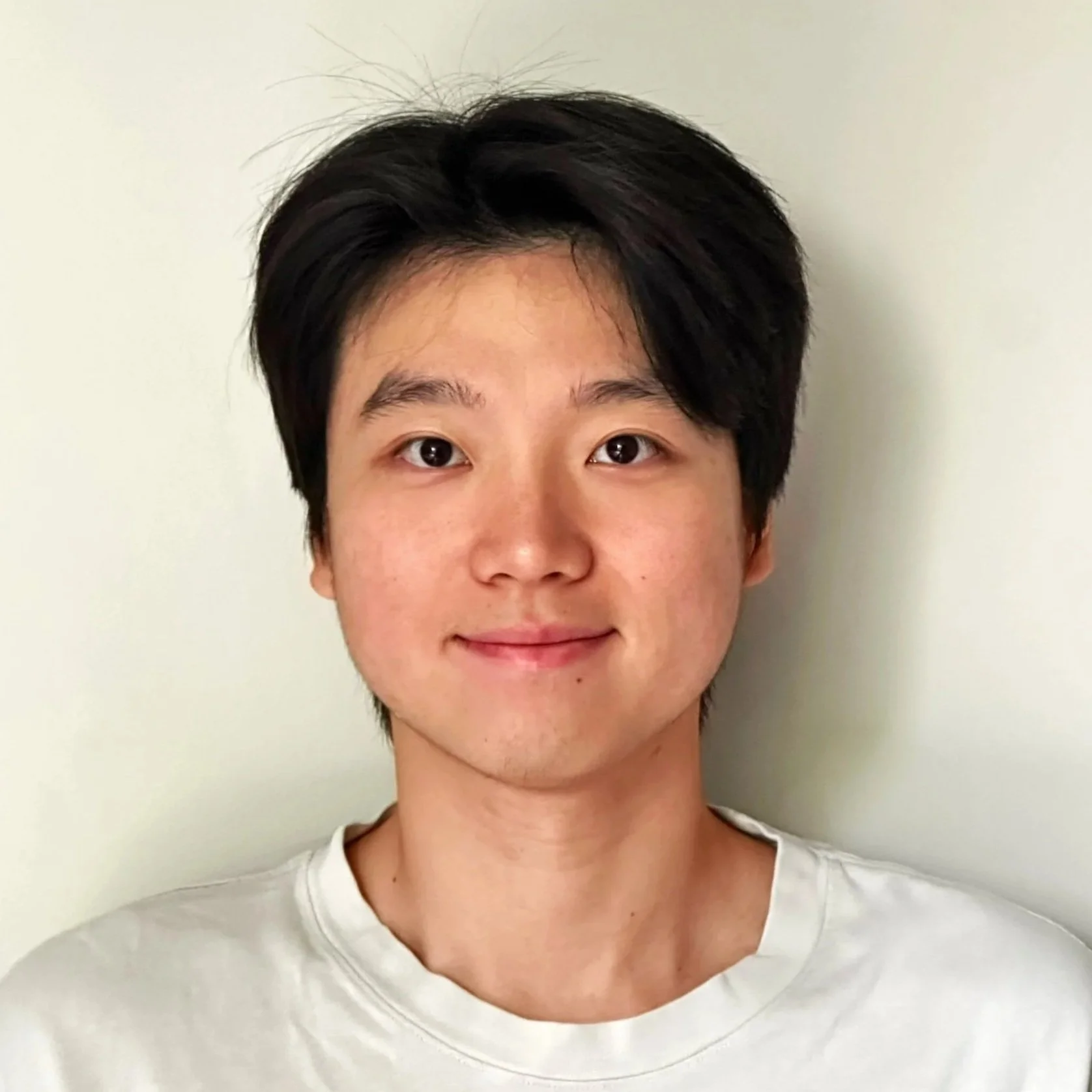 A young man with dark hair, smiling softly, wearing a white t-shirt, standing against a plain off-white wall.