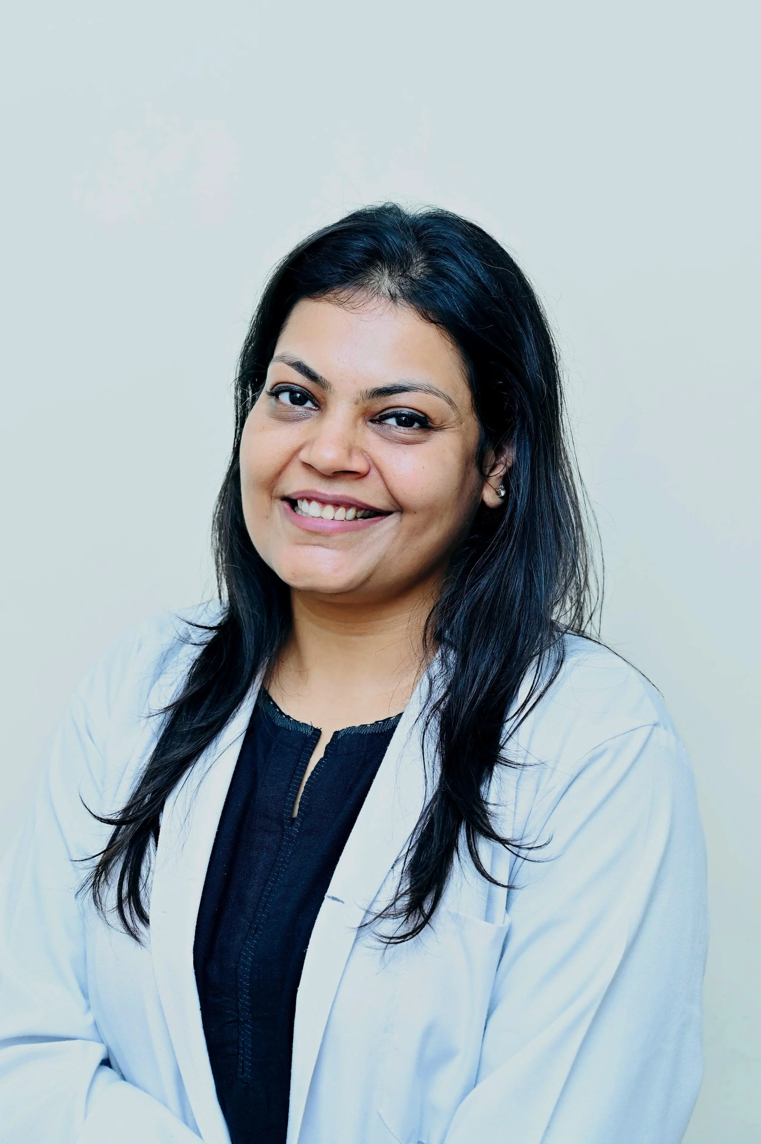 A woman with long dark hair, smiling, wearing a white lab coat over a black shirt, standing against a plain white background.