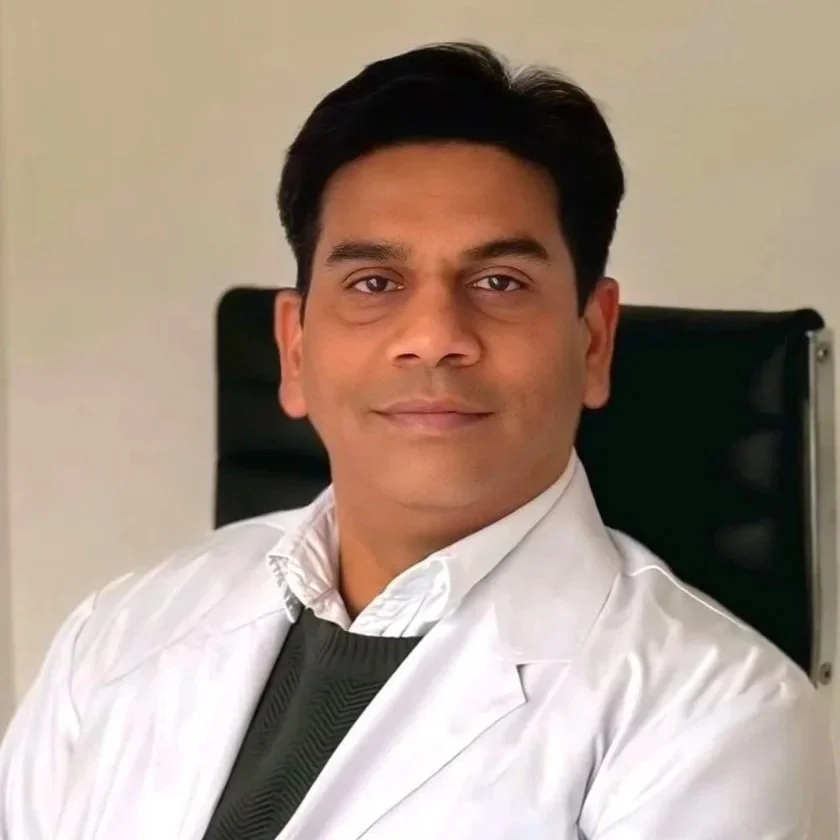 A man wearing a white medical coat, sitting in an office chair, smiling at the camera.
