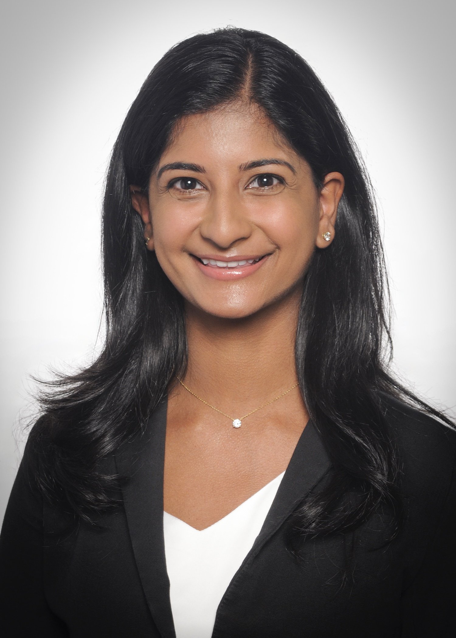 Professional portrait of a woman with long black hair, wearing a black blazer over a white top, gold jewelry, and smiling against a light gray background.