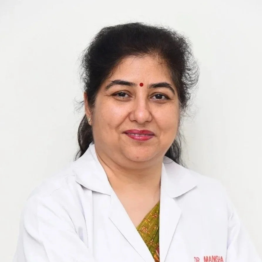 Portrait of a woman with black hair, wearing a white coat with a red dot bindi on her forehead, smiling, against a plain white background.