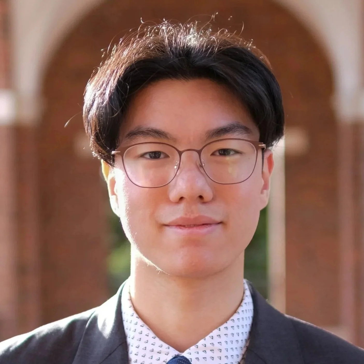 A young man with black hair and glasses wearing a suit and patterned shirt outdoors with a blurred architectural background.